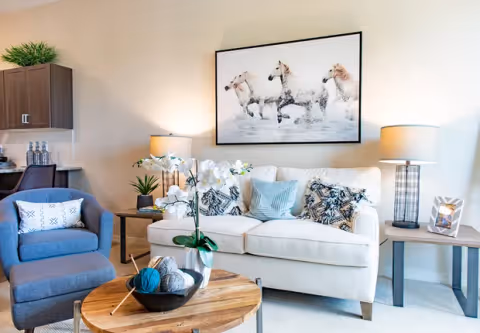 A cozy living room with a white sofa adorned with patterned and solid pillows, a blue armchair with a white pillow, a round wooden coffee table with a bowl of yarn and knitting needles, and two side tables with lamps and decorative items. A large framed picture of three white horses running through water hangs on the wall above the sofa.