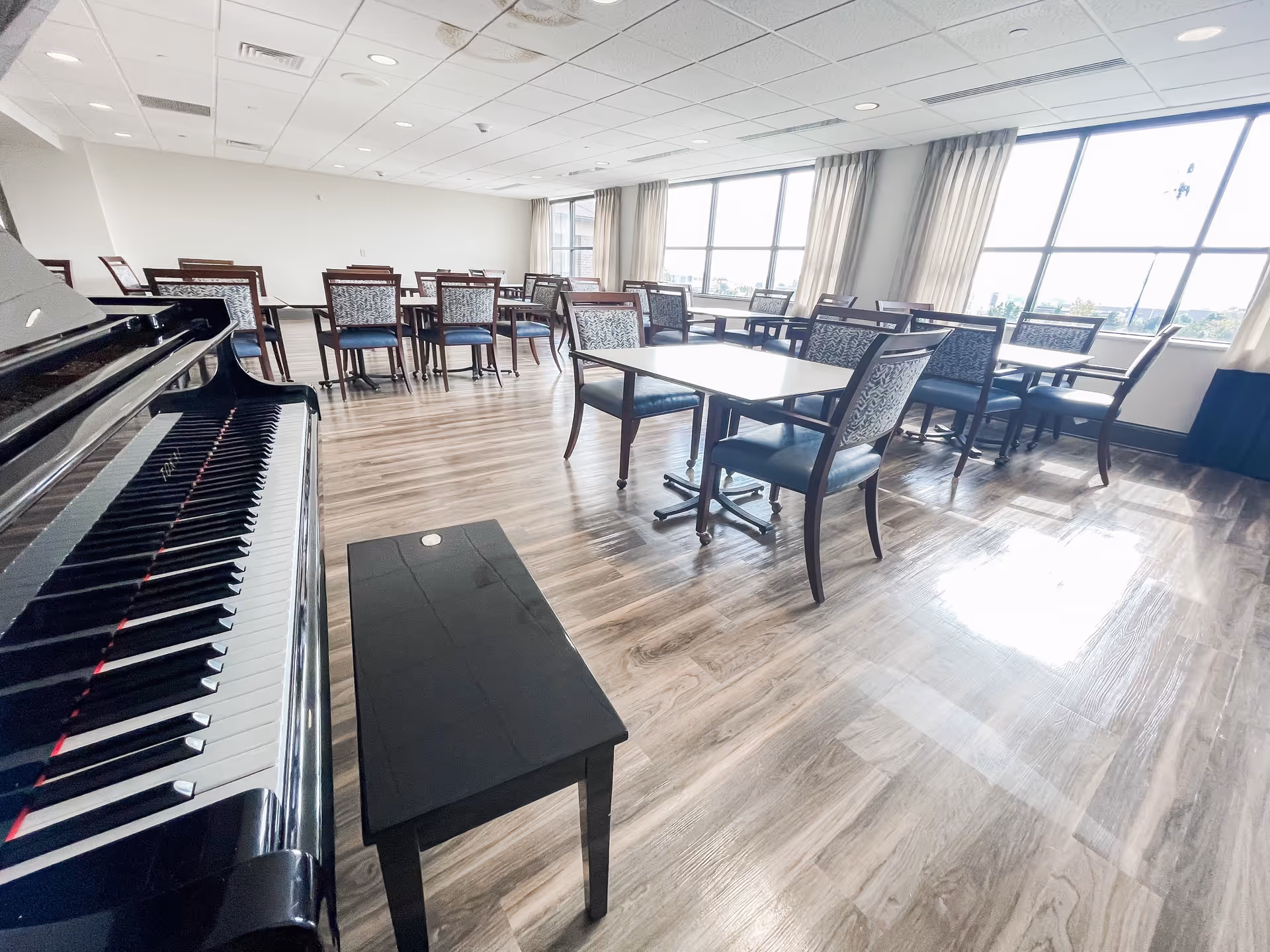 A bright room with large windows letting in natural light, featuring multiple tables and chairs arranged in a dining area. A black grand piano with a matching bench is positioned in the foreground on the left side. The floor is covered with wood-patterned flooring, and the ceiling has recessed lighting and air vents.