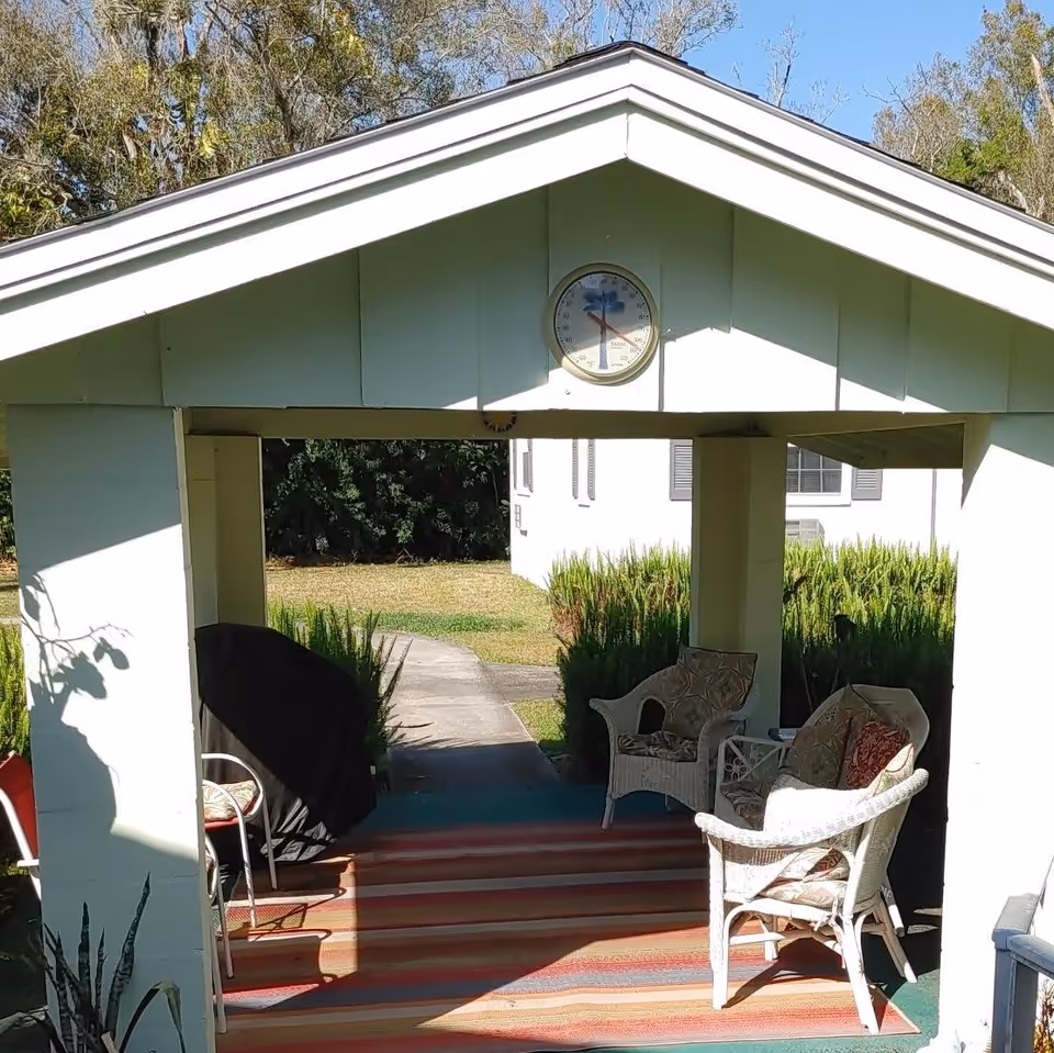 Outdoor covered patio area with a striped rug, white wicker chairs with cushions, a small table, and a covered grill. A clock is mounted on the wall above the entrance. Green bushes and a white building are visible in the background under a clear blue sky.