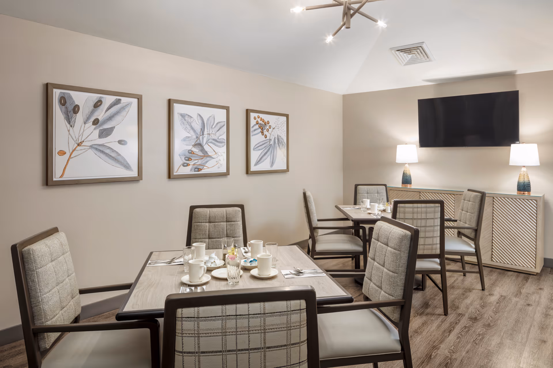 A dining room with two tables set for meals, each surrounded by four cushioned chairs. The tables have cups, glasses, plates, and utensils arranged neatly. On the wall are three framed botanical prints. A flat-screen TV is mounted above a sideboard with two table lamps on either side. The room has wood flooring and neutral-colored walls.