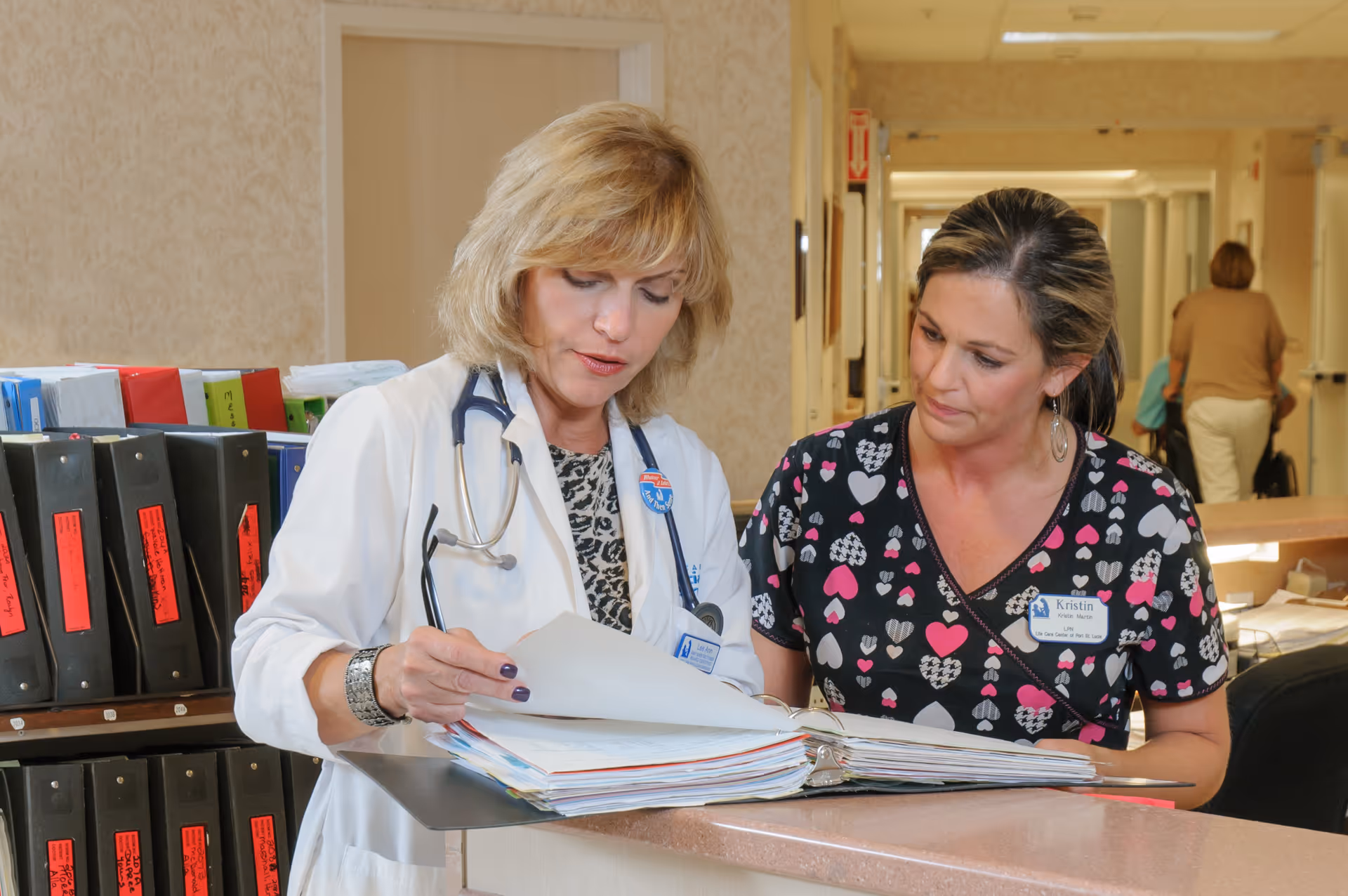 Two female healthcare workers reviewing documents at a reception desk in a medical or senior care facility. One is wearing a white coat with a stethoscope, and the other is in a patterned scrub top. There are binders and files on the desk and shelves behind them, with a hallway in the background where another person is walking away.
