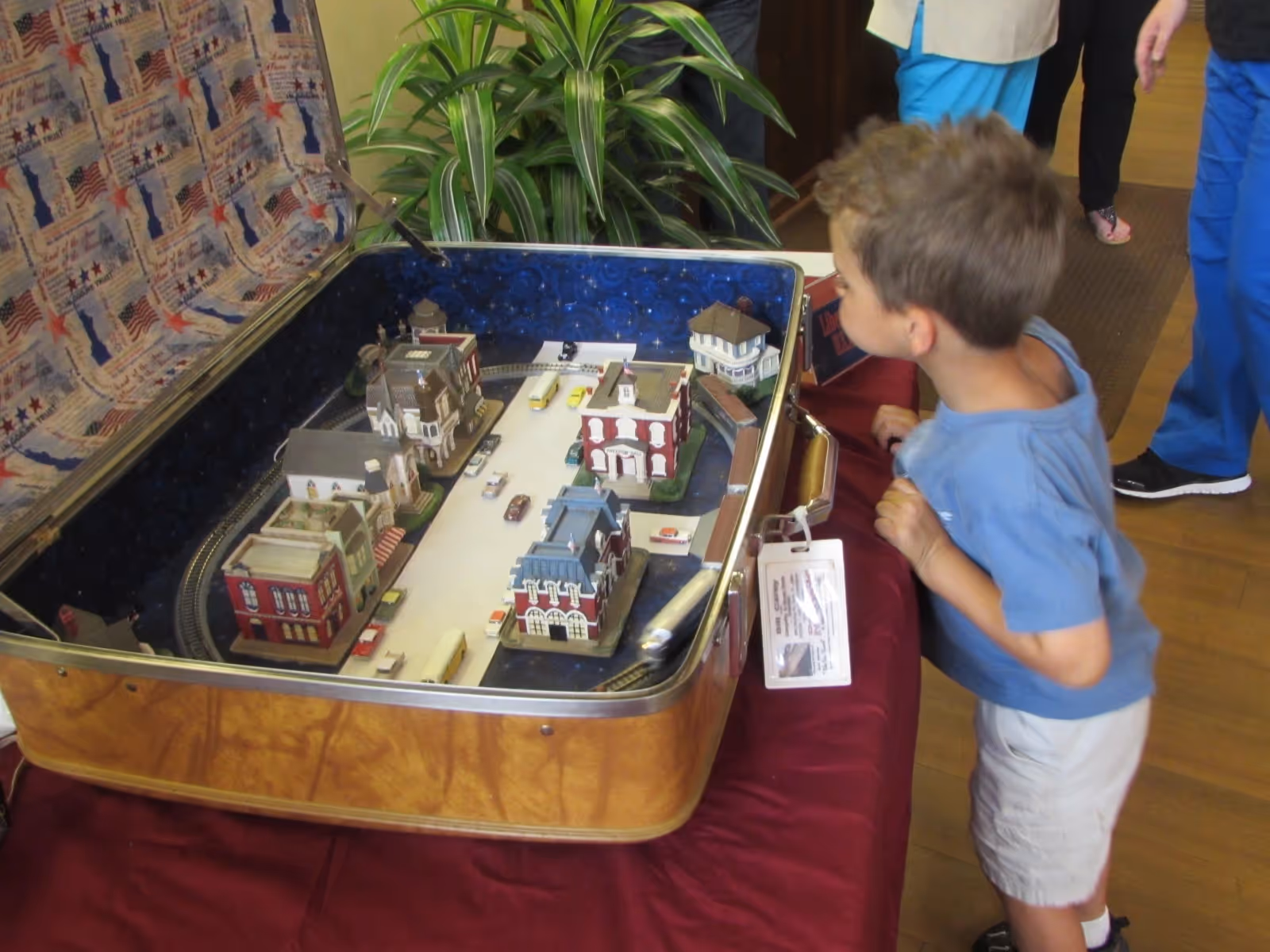 A young boy leans over a table to look at a miniature model town displayed inside an open suitcase.