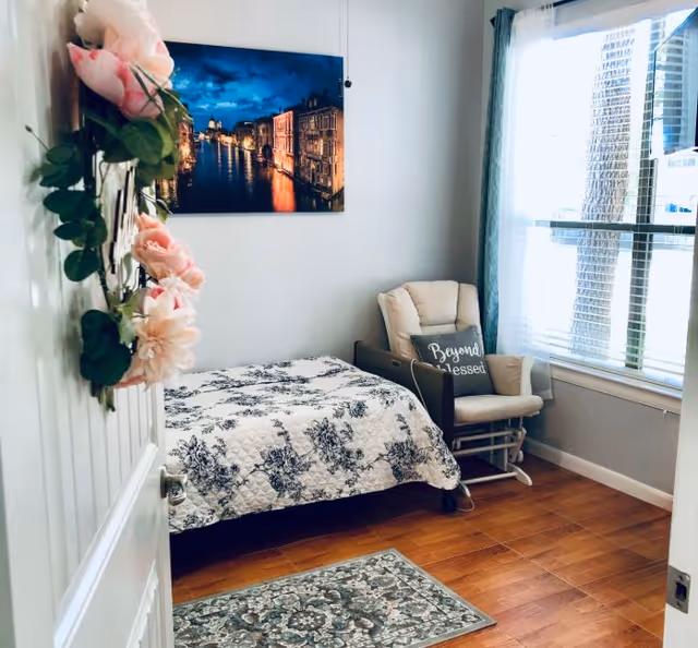 A cozy bedroom with a single bed covered in a white and black floral quilt. A cushioned rocking chair with a pillow that says 'Beyond Blessed' is placed near a large window with blue curtains. A decorative floral wreath hangs on the door, and a framed picture of a nighttime cityscape over water is mounted on the wall. The floor is wooden with a patterned rug near the bed.