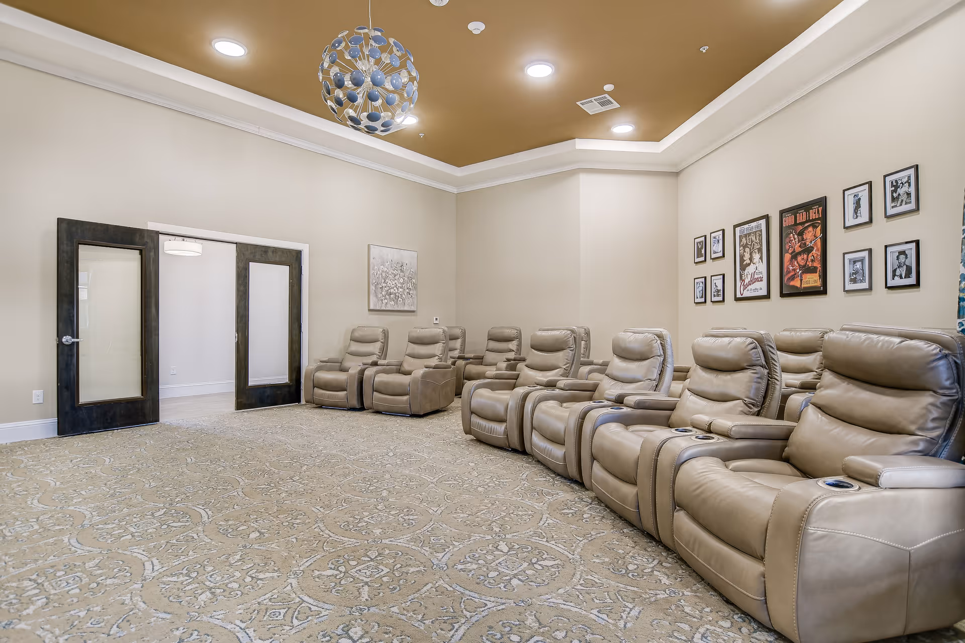A senior living facility lounge with a row of beige leather recliner chairs facing a blank wall. The room has a patterned carpet, beige walls, and a ceiling with recessed lighting and a decorative light fixture. There are framed pictures and posters on one wall and double glass doors on the opposite side.