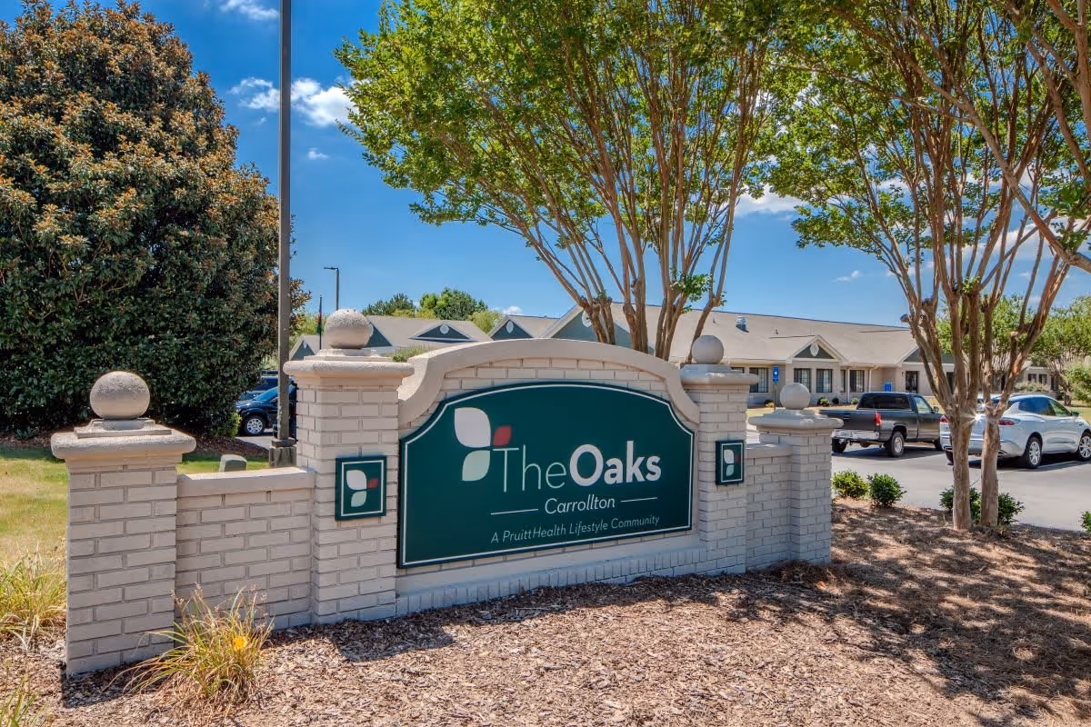 Outdoor view of the entrance sign for The Oaks Carrollton, a PruittHealth Lifestyle Community, with a brick structure surrounded by trees and parked cars in the background under a blue sky.