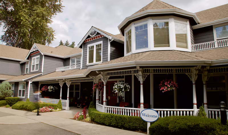 Exterior view of a large, two-story senior living facility building with gray siding and white trim. The building features a covered porch with decorative columns and hanging flower baskets. A small sign in front of the porch reads 'Welcome'. There are bushes and landscaped plants along the walkway leading to the entrance.