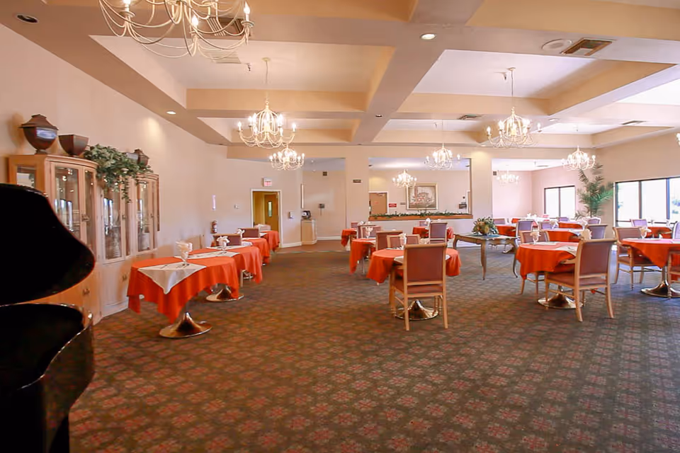 A spacious dining room with multiple round tables covered with red tablecloths and white napkins. The room features chandeliers hanging from the ceiling, a patterned carpet, large windows letting in natural light, a wooden cabinet with decorative items, and a piano on the left side.