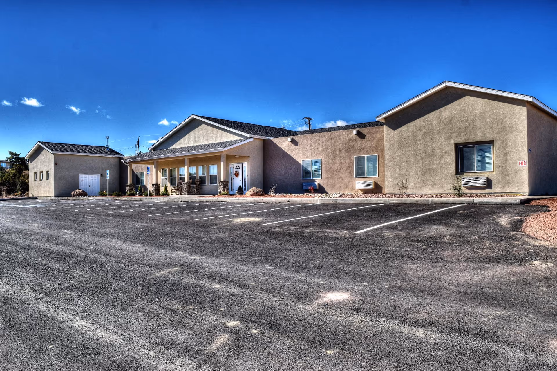 Exterior view of a single-story beige building with a covered entrance and several windows, situated under a clear blue sky with a large empty parking lot in front.