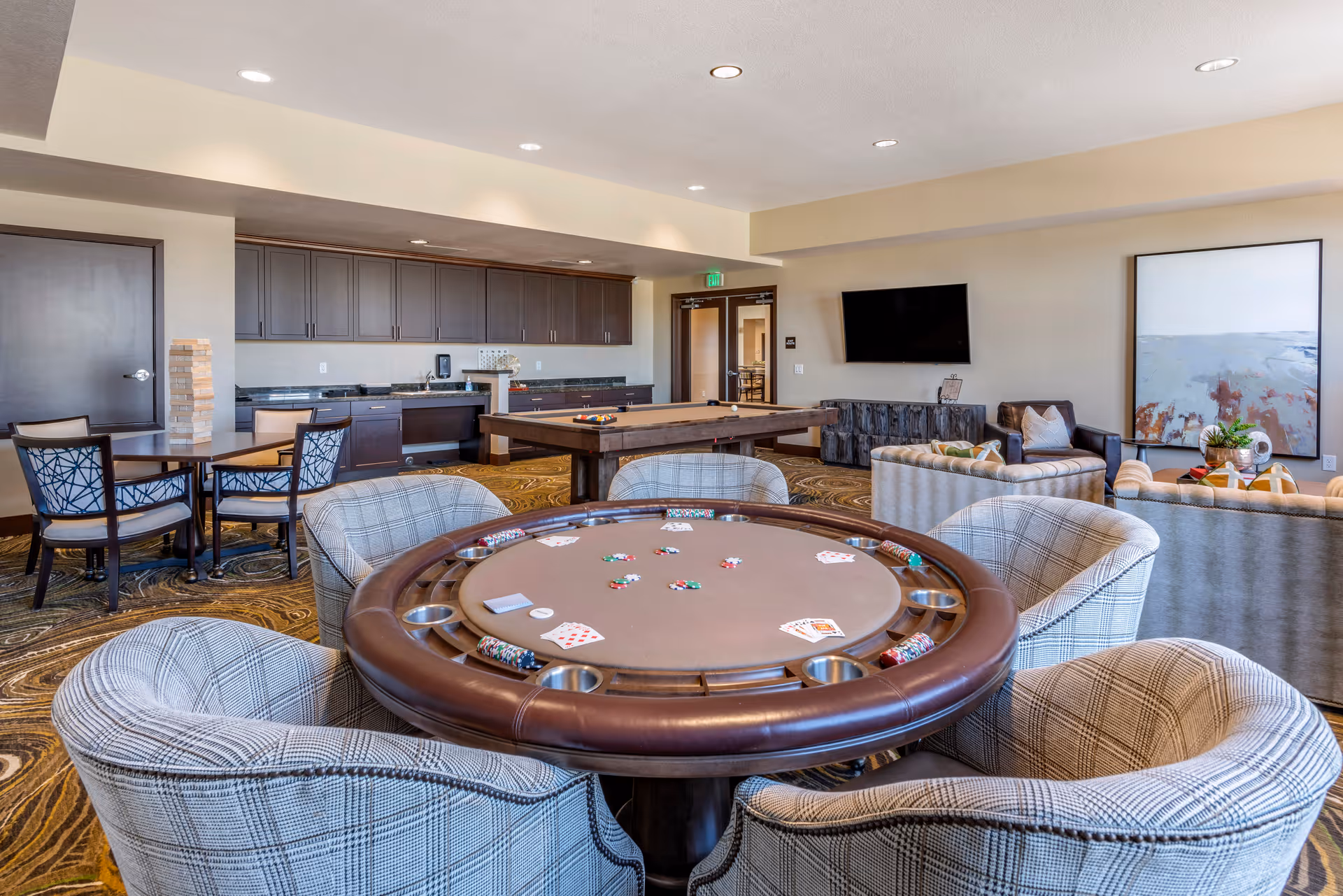 A senior living community lounge with a poker table and cushioned chairs, a pool table, kitchenette, seating area and wall-mounted TV.