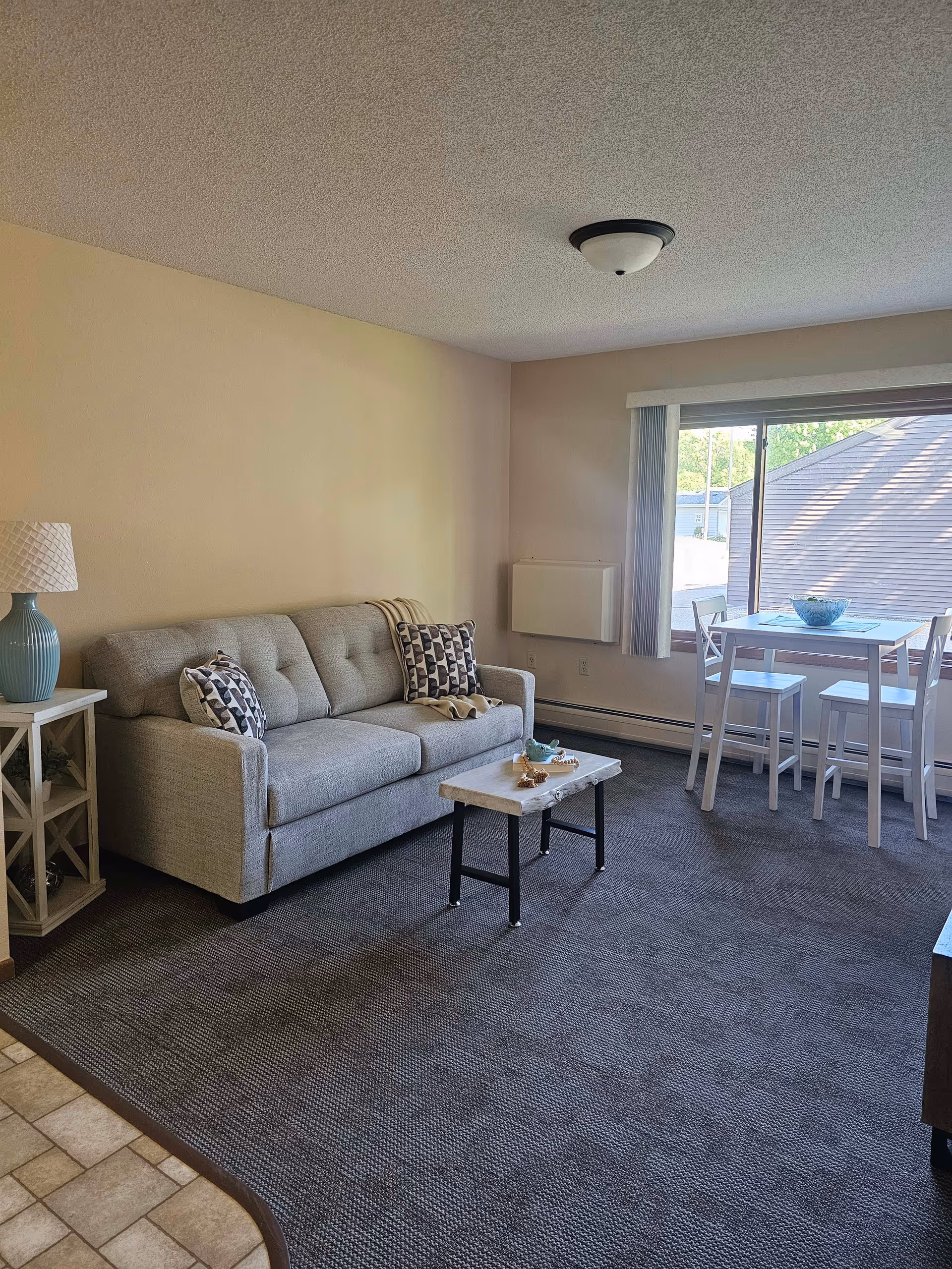 A cozy living room with a light gray sofa adorned with two patterned pillows and a beige throw blanket. Next to the sofa is a small white side table with a blue lamp. In front of the sofa is a small coffee table with decorative items. A large window with vertical blinds lets in natural light, and near the window is a small white dining table with four chairs. The room has beige walls and a dark carpeted floor.