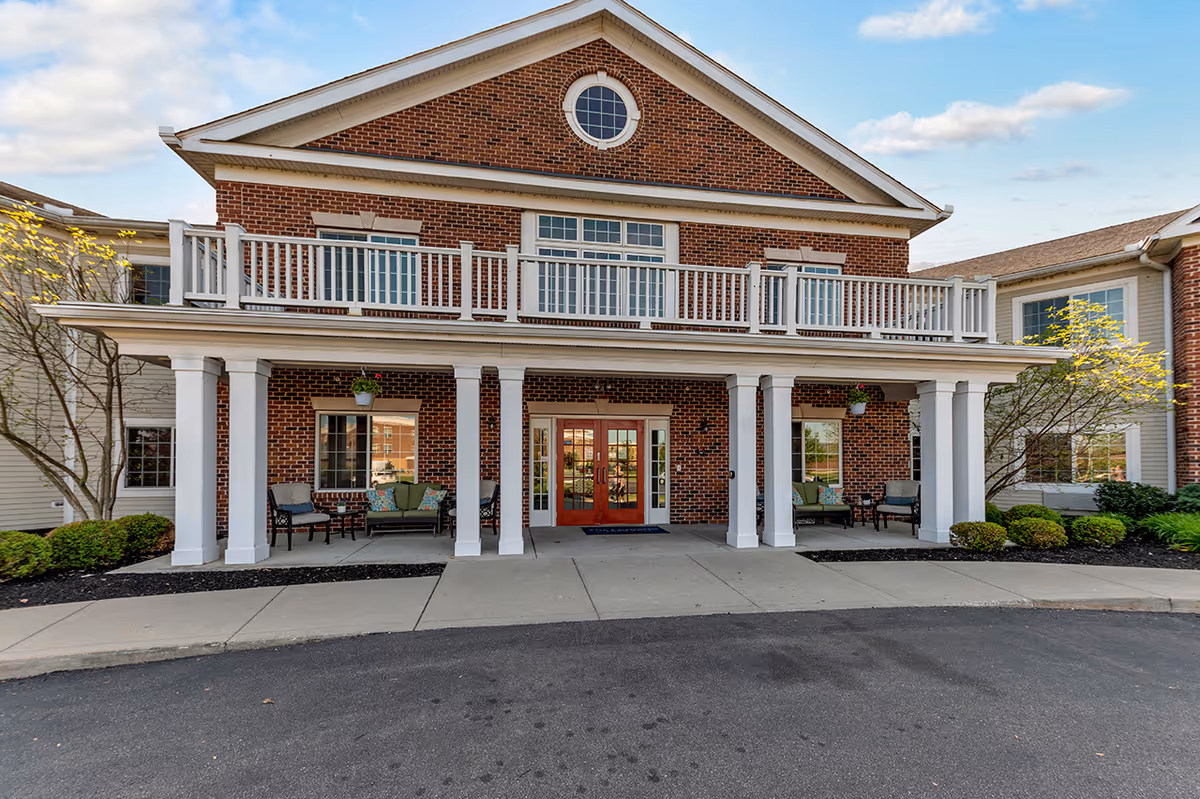 Front entrance of a two-story brick building with white columns, a covered porch, balcony railings, and outdoor seating.