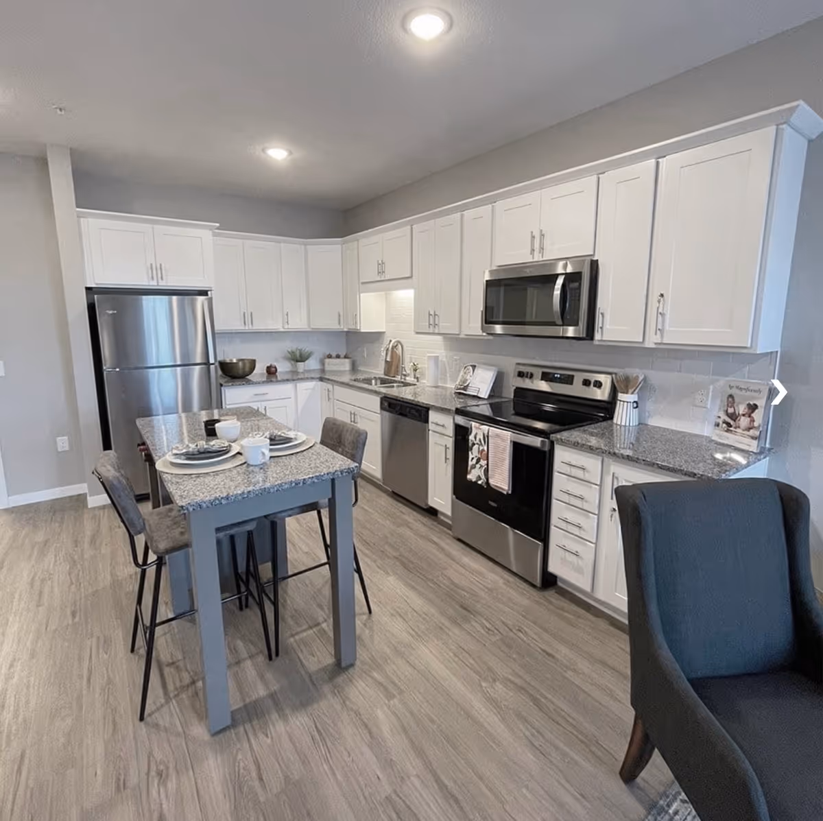 Modern kitchen with white cabinets, stainless steel appliances including a refrigerator, dishwasher, stove, and microwave. A small dining table with two chairs is set with plates, cups, and napkins. The floor is light wood, and there is a dark upholstered chair partially visible on the right.