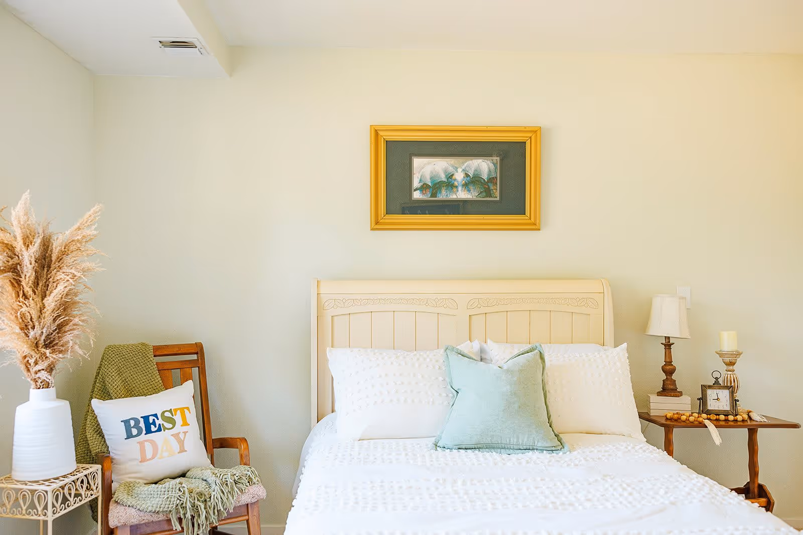 A cozy bedroom with a cream-colored bed featuring white and light green pillows. To the left, there is a wooden chair with a green throw blanket and a pillow that says 'BEST DAY'. A white vase with dried pampas grass sits on a small table next to the chair. On the right side of the bed, there is a wooden nightstand with a lamp, a candle, a clock, and some decorative items. A framed picture with a gold frame hangs on the wall above the bed.