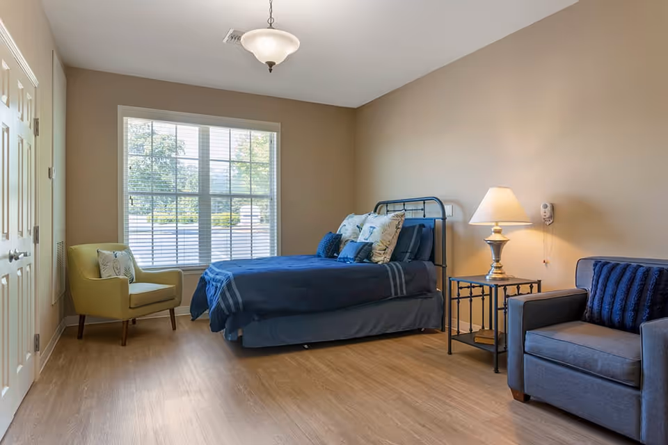 A cozy bedroom with a blue bed, multiple pillows, a yellow armchair near a large window with blinds, a side table with a lamp, and a gray armchair with a blue cushion. The room has wooden flooring and beige walls.