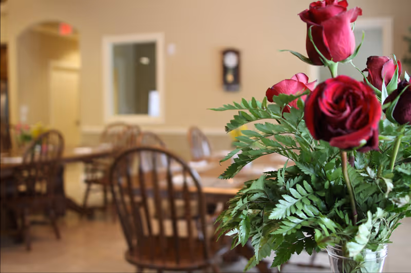 Close-up of a bouquet of red roses and green foliage in a glass vase with a dining area in the background featuring wooden chairs and tables in a softly lit room.
