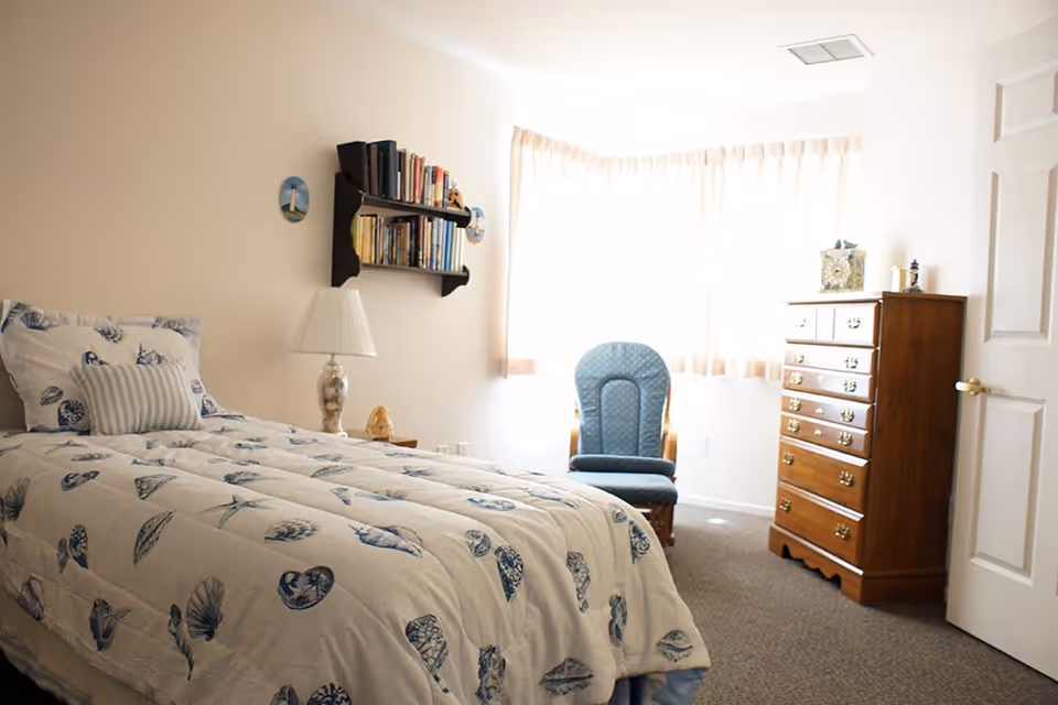 A bright bedroom with a single bed covered in a seashell-patterned comforter and matching pillow. A wooden shelf with books is mounted on the wall above a bedside table with a lamp. A blue cushioned chair is placed near a window with sheer curtains, and a tall wooden dresser stands against the wall next to a partially open door.