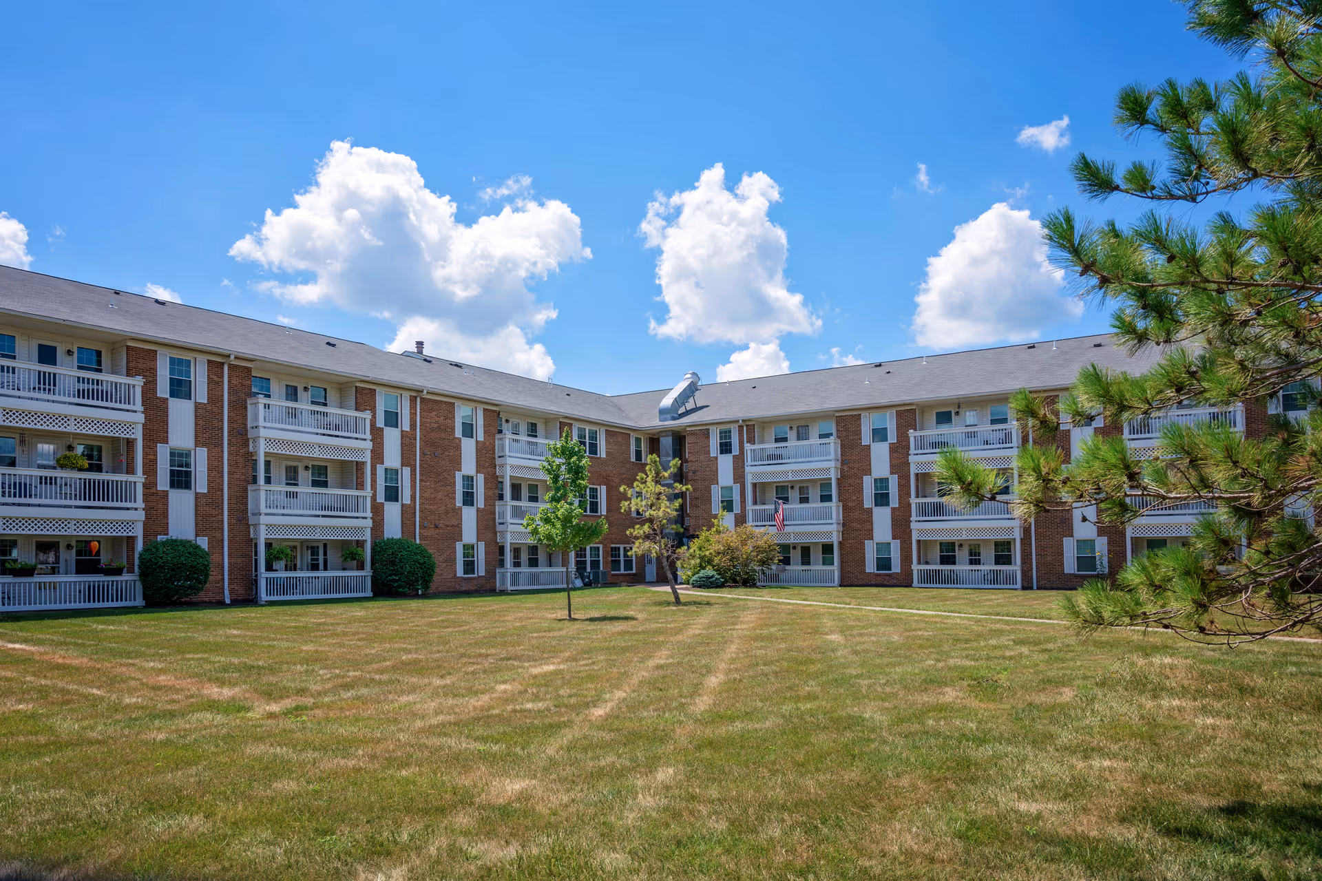 Exterior view of a three-story senior living facility building with balconies, surrounded by a large grassy lawn and a few trees under a blue sky with scattered clouds.