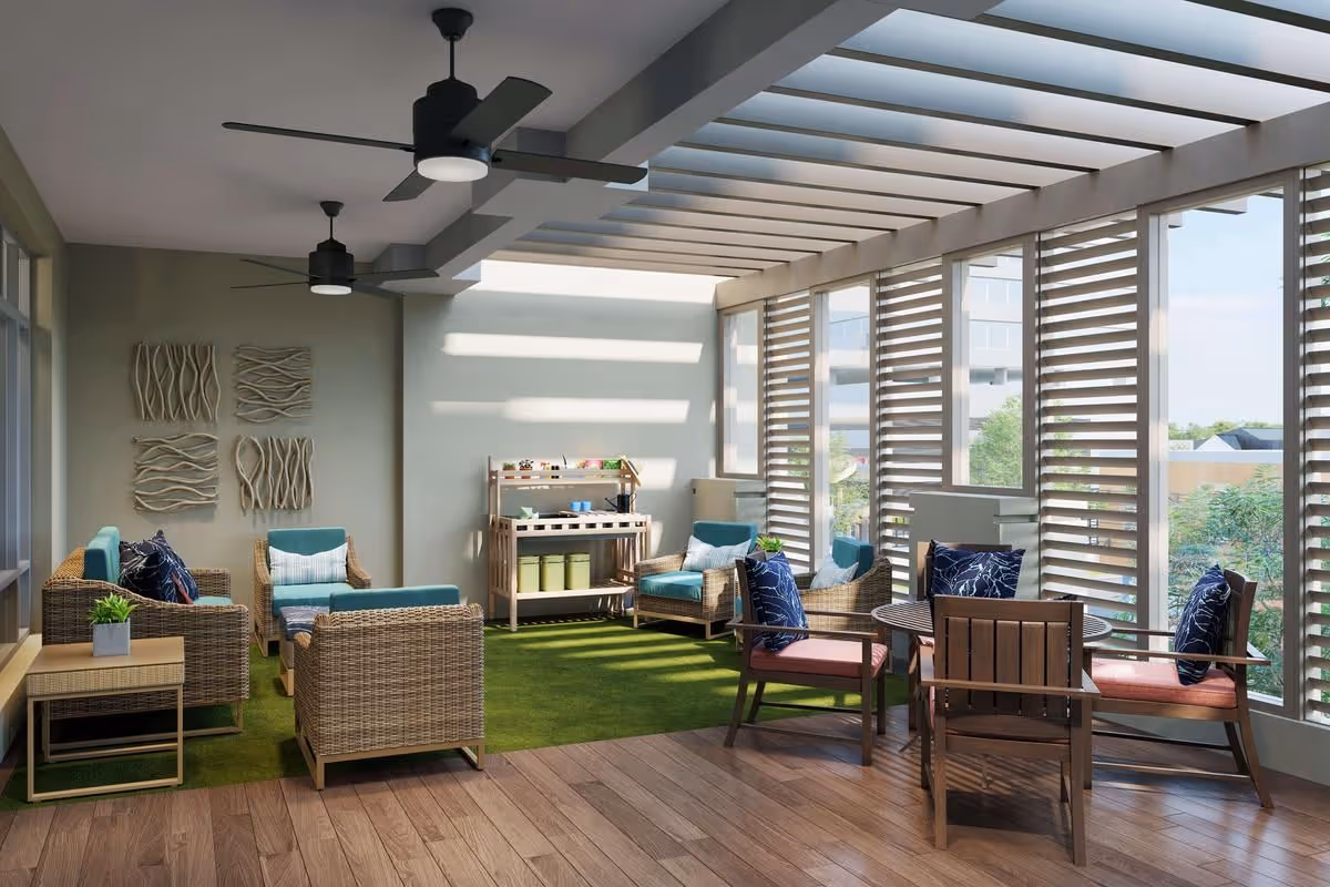 A bright indoor lounge area with large windows and slatted shades allowing natural light to enter. The space features wicker armchairs and sofas with teal cushions, a small wooden table with potted plants, and a round wooden table with chairs that have dark blue patterned pillows. The floor is a combination of wood and green carpet, and the ceiling has modern black ceiling fans with lights.