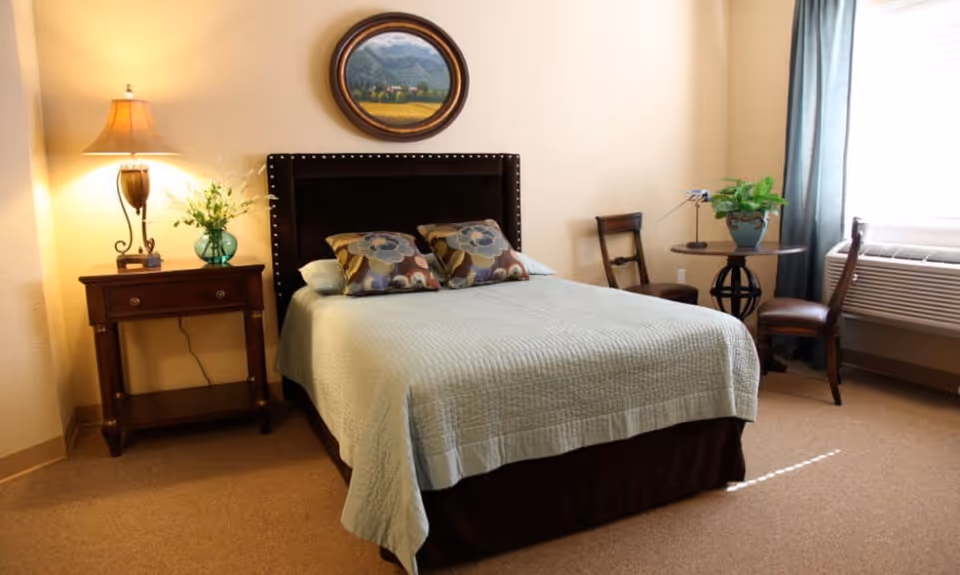 A cozy bedroom with a neatly made bed featuring a light blue quilt and two floral pillows. To the left of the bed is a wooden nightstand with a lamp and a vase of flowers. Above the bed hangs a round framed painting of a landscape. To the right, there is a small round table with a potted plant and two wooden chairs near a window with blue curtains and an air conditioning unit below it.