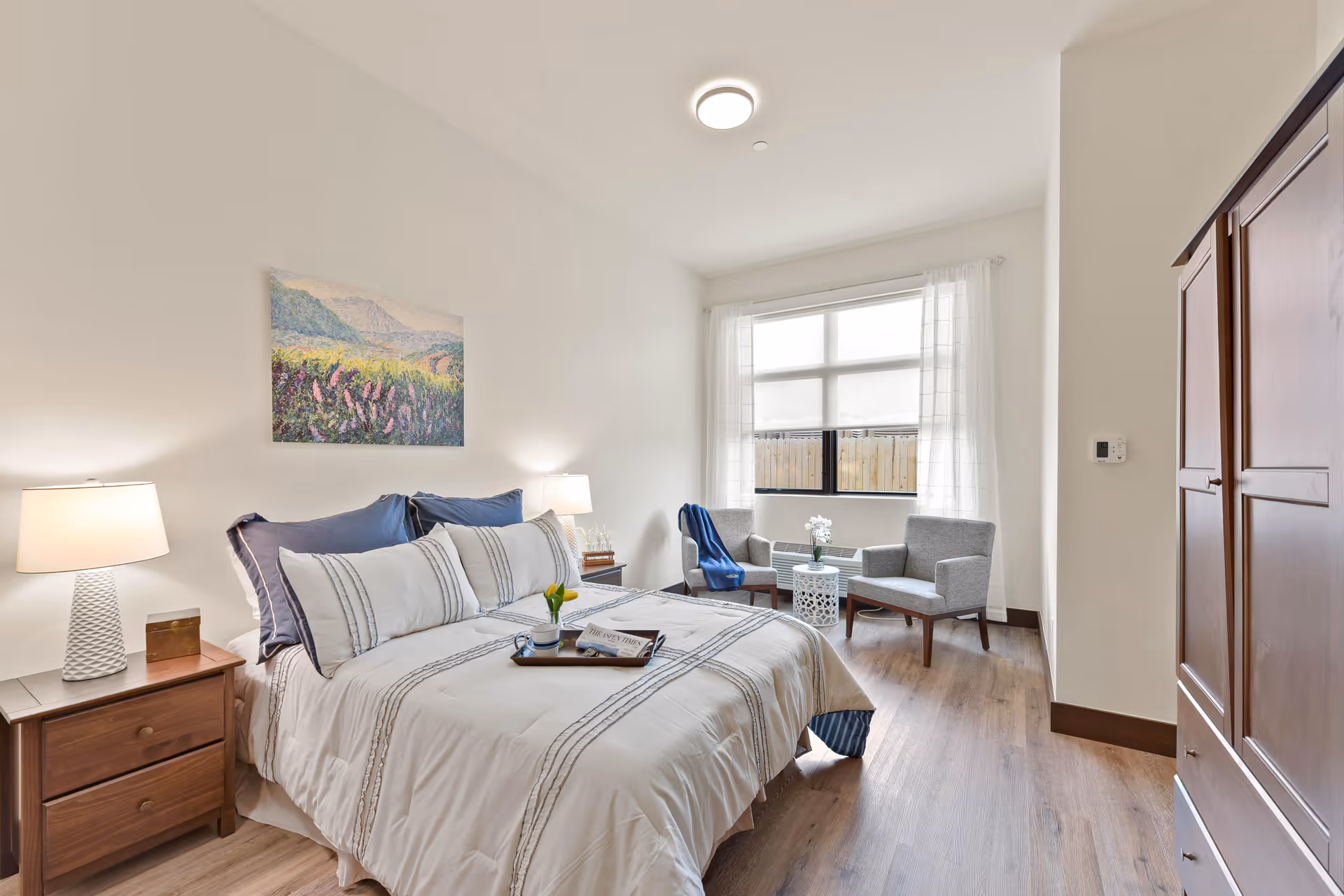 Well-lit bedroom with a made bed, bedside tables and lamps, a seating area by a window, and a wooden armoire.