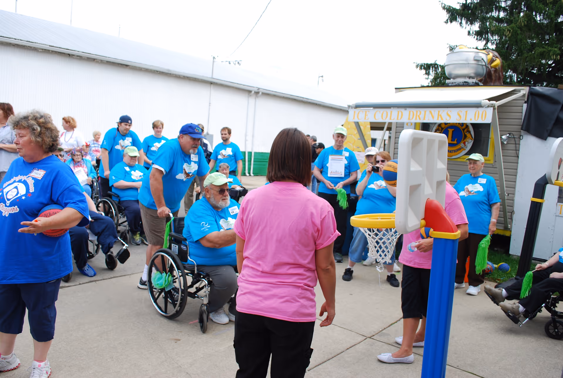 A group of elderly people and caregivers participating in an outdoor activity. Several individuals are in wheelchairs, and many are wearing matching blue t-shirts. A woman in a pink shirt stands near a small basketball hoop, while others watch and engage in the event. A sign in the background advertises ice cold drinks for $1.00.