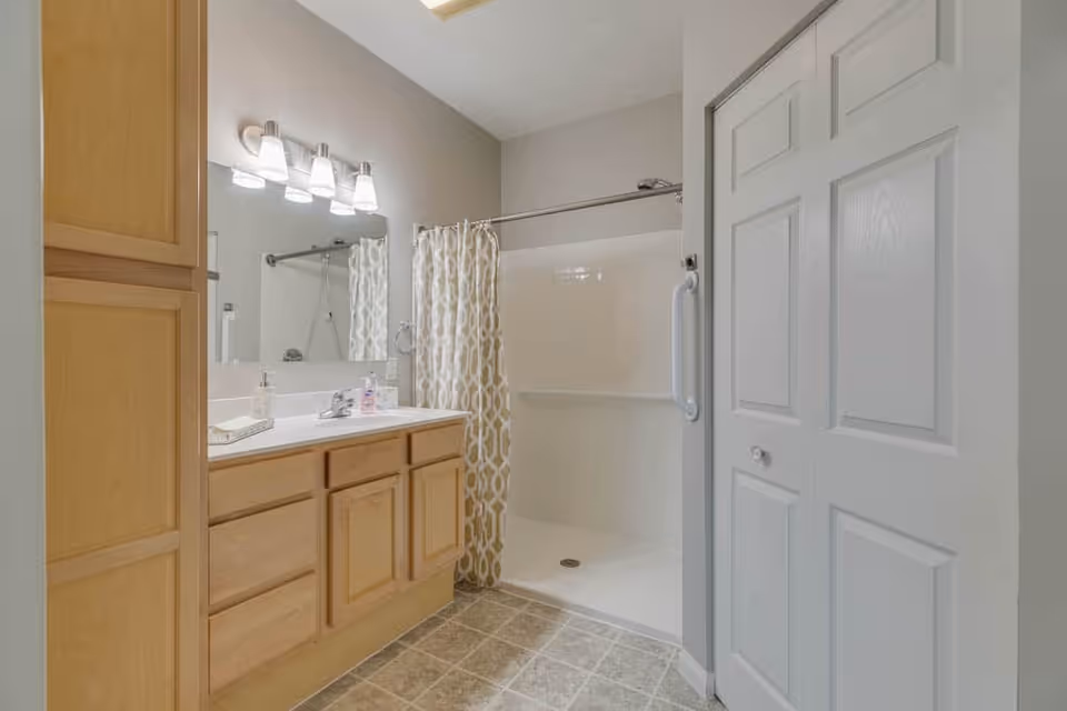 A clean and well-lit bathroom featuring a wooden vanity with a white countertop, a large mirror with four light fixtures above it, a walk-in shower with a patterned shower curtain, and a white door with a grab bar attached to the wall beside it.