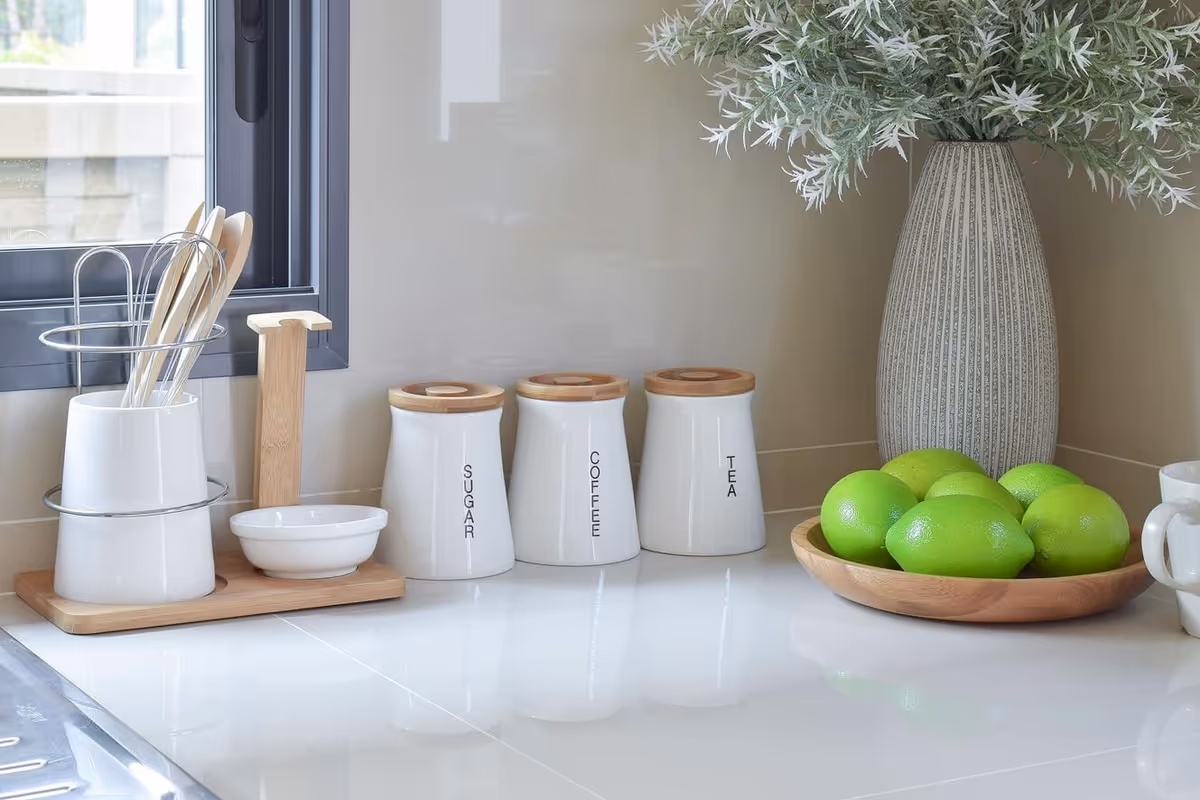 Bright kitchen countertop featuring utensil holders, three labeled ceramic canisters (Sugar, Coffee, Tea), a vase with greenery, and a wooden bowl of green limes.