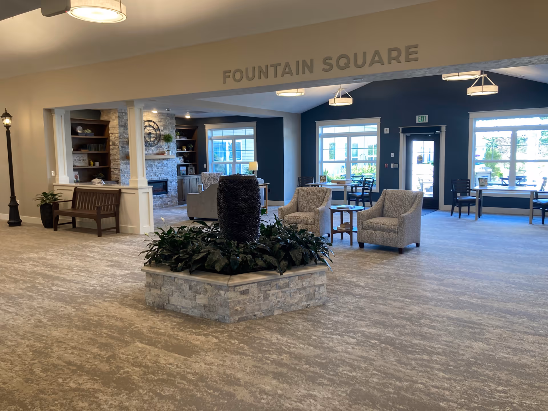 A spacious and well-lit common area named Fountain Square in a retirement community. The room features a central decorative water fountain surrounded by plants, comfortable armchairs with a small table, large windows letting in natural light, a stone fireplace with built-in shelves, and additional seating including benches and tables with chairs.