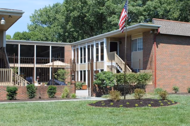 Exterior view of a brick building with large windows and a screened porch area, surrounded by green grass, shrubs, and trees. An American flag is mounted on a flagpole in front of the building under a clear blue sky.