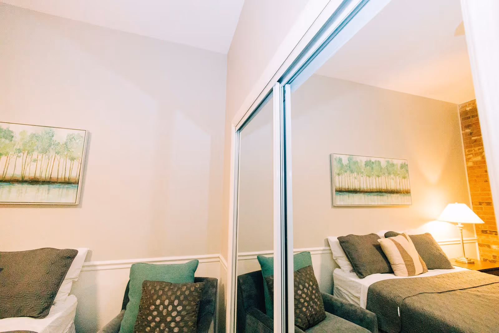 A cozy bedroom with a bed featuring multiple pillows and a green throw blanket. There is a green armchair with two cushions next to the bed. A framed painting of trees hangs on the wall above the bed. A bedside table with a lamp is visible, and a mirrored closet door reflects part of the room.