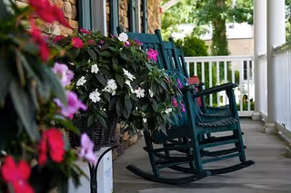 A porch with dark green rocking chairs lined up along a stone wall. There are flower baskets with pink, white, and red flowers hanging on the railing and placed near the chairs. The porch has a white railing and overlooks a green outdoor area with trees.