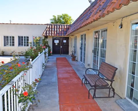 Outdoor walkway along a beige building with a red path, bench, and flowering railing.