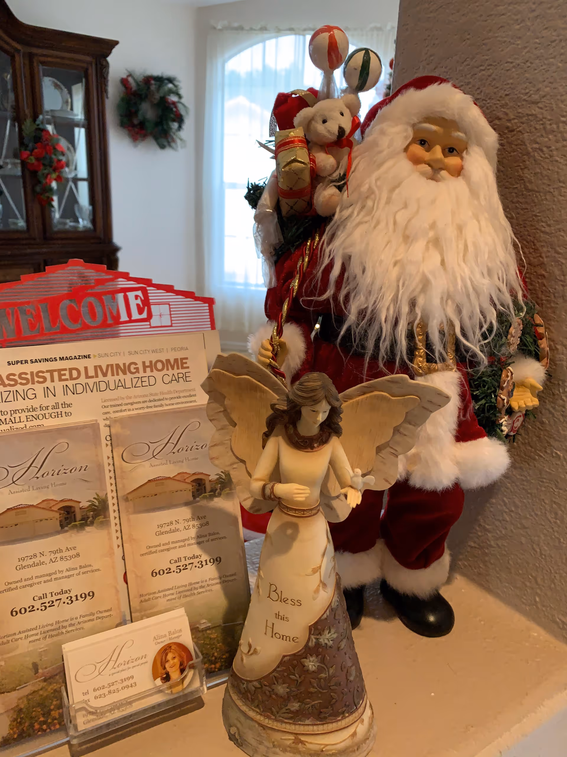 A decorative display featuring a Santa Claus figurine holding gifts and a teddy bear, an angel figurine with the words 'Bless this Home' on its dress, and brochures for Horizon Assisted Living Home LLC. In the background, there is a wooden cabinet with glass doors and a window with sheer curtains.