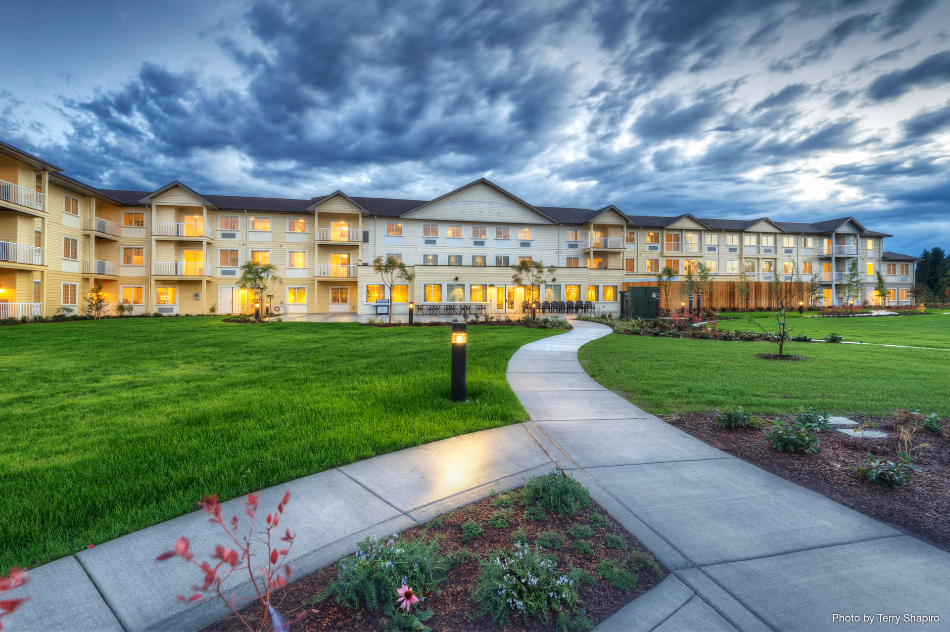 Exterior view of a large, three-story senior living facility building with multiple balconies and windows illuminated from inside during dusk. A curved concrete pathway leads through a well-maintained green lawn and landscaped garden beds towards the entrance. The sky is cloudy with dramatic lighting.