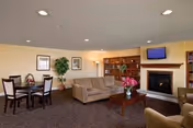 A cozy living room area with a beige sofa, armchair, wooden coffee table with a vase of pink flowers, a fireplace with a mounted TV above it, a round dining table with four chairs, framed pictures on the walls, a potted plant, and built-in wooden shelves.