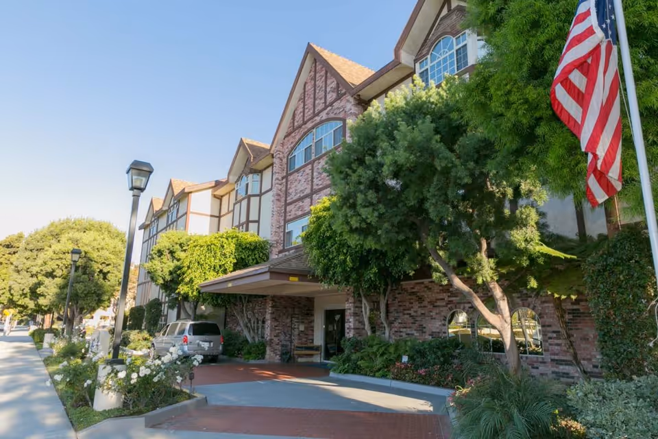 Exterior view of a multi-story assisted living facility with brick and timber facade, surrounded by trees and greenery. An American flag is visible on the right side near the entrance, and a vehicle is parked under a covered driveway.