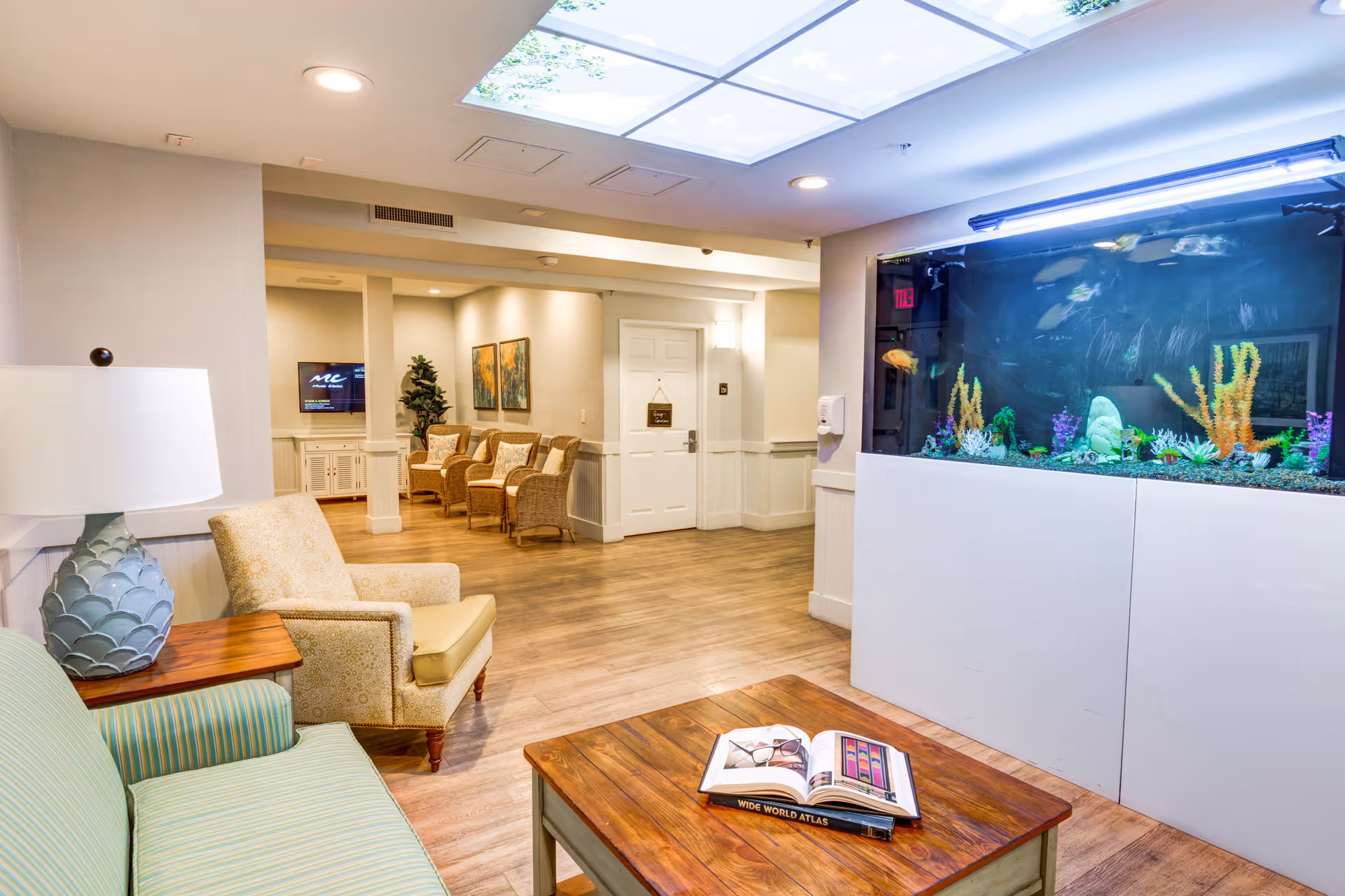 A bright and welcoming interior lounge area with a green striped sofa, a beige armchair, a wooden coffee table with an open book and glasses on it, and a large built-in aquarium filled with colorful fish and aquatic plants. In the background, there are additional wicker chairs, a TV mounted on the wall, and framed artwork. The ceiling features a light panel with a sky and tree design.