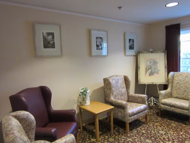 A cozy sitting area with four armchairs arranged around a small wooden side table holding a vase with white flowers. The walls are decorated with three framed black and white pictures and a framed painting on an easel near a window with dark curtains. The floor is covered with a patterned carpet.