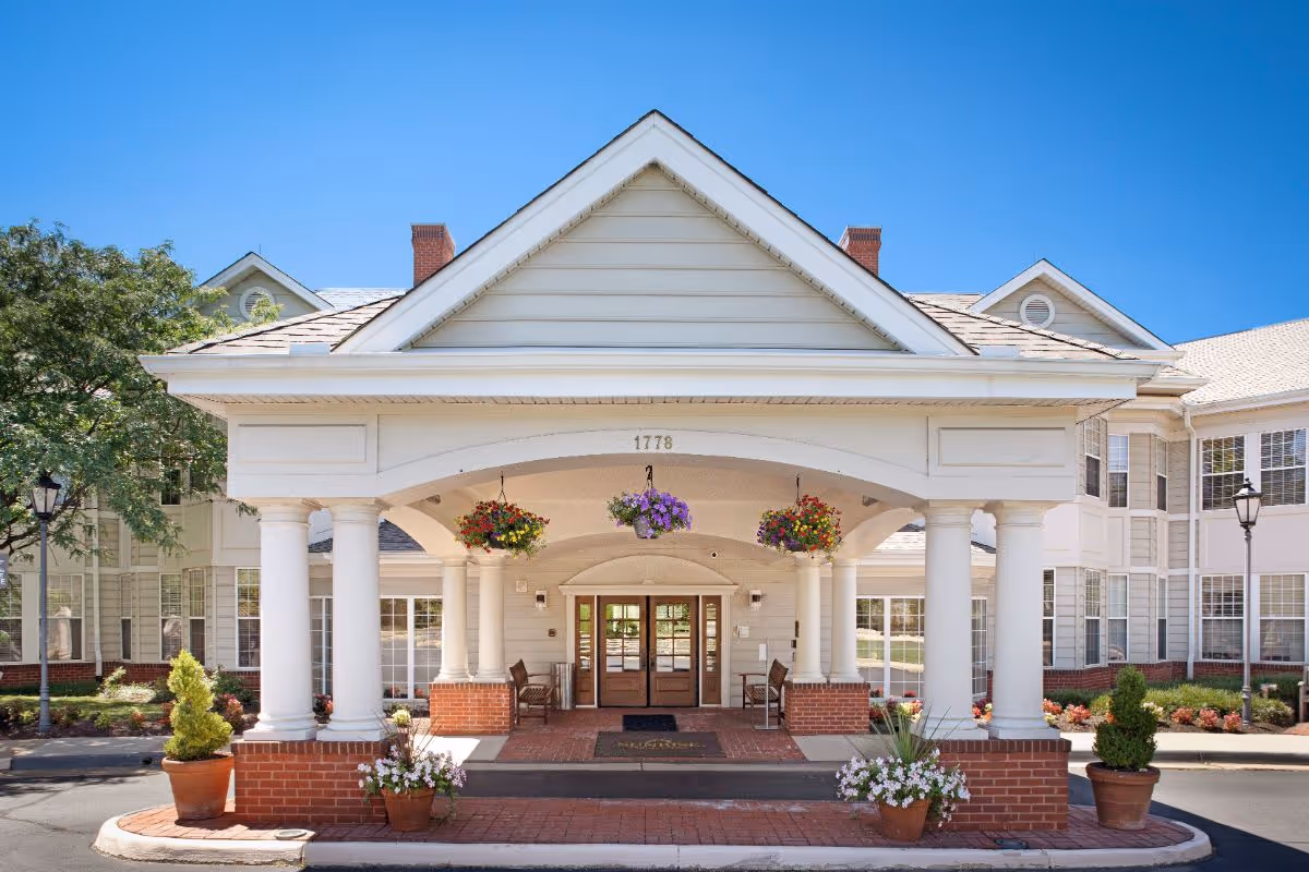 Front exterior view of Sunrise at Reston Town Center senior living facility featuring a covered entrance with white columns, hanging flower baskets, potted plants, and double glass doors under a clear blue sky.