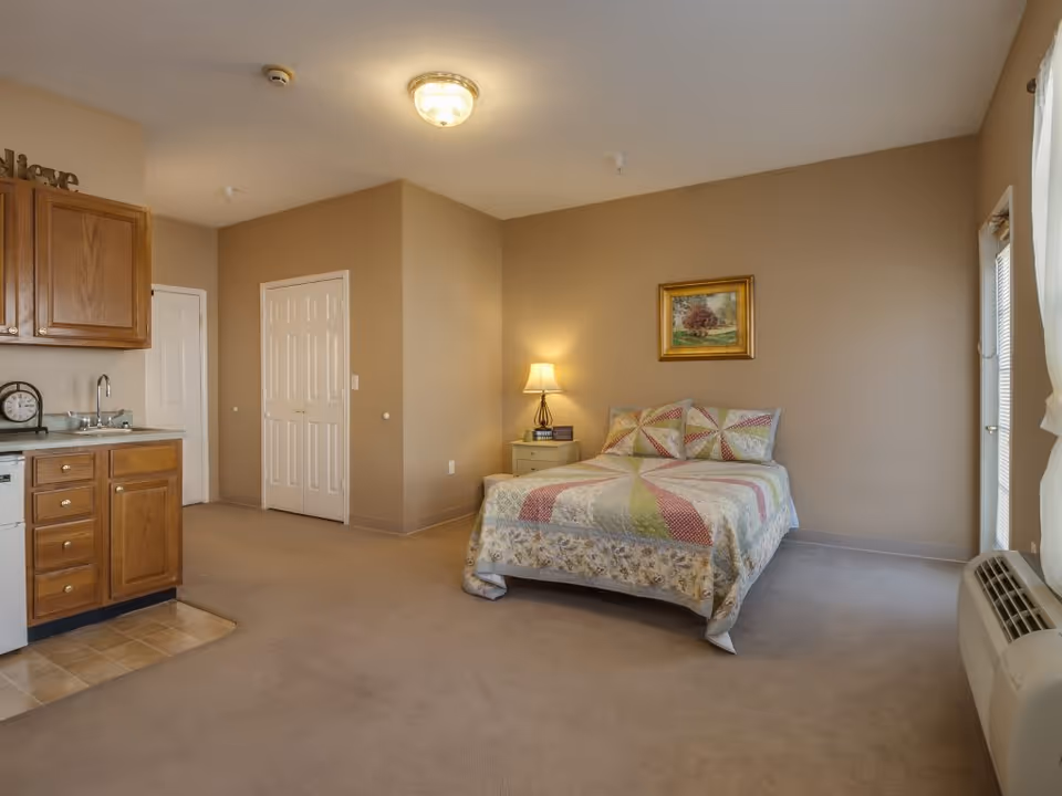A cozy senior living studio apartment featuring a bed with a colorful quilt, a nightstand with a lamp, a framed painting on the wall, a small kitchenette with wooden cabinets and a sink, and a window with curtains letting in natural light.