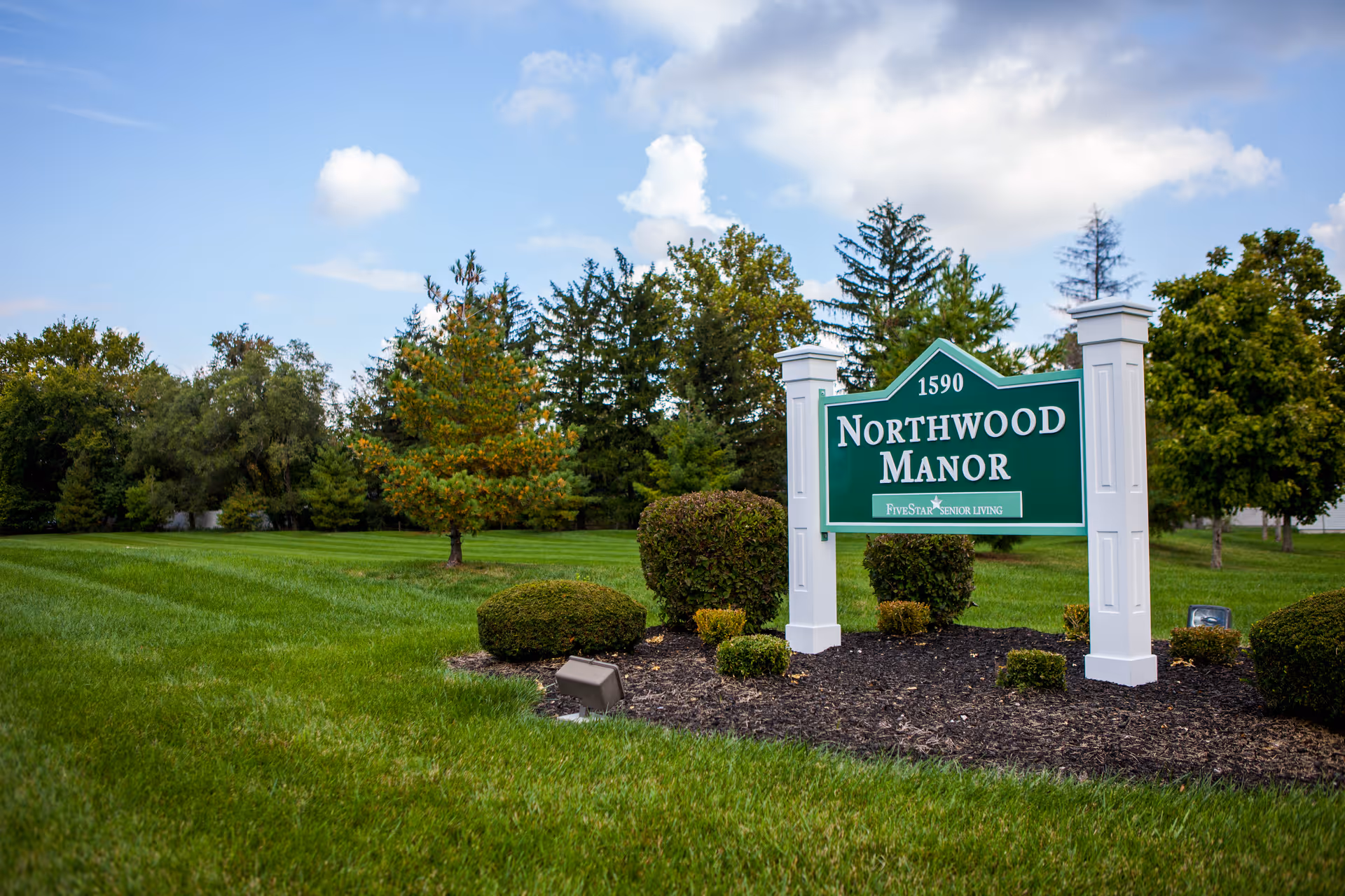 A green Northwood Manor sign on white posts set in landscaped grass and shrubs with trees in the background.