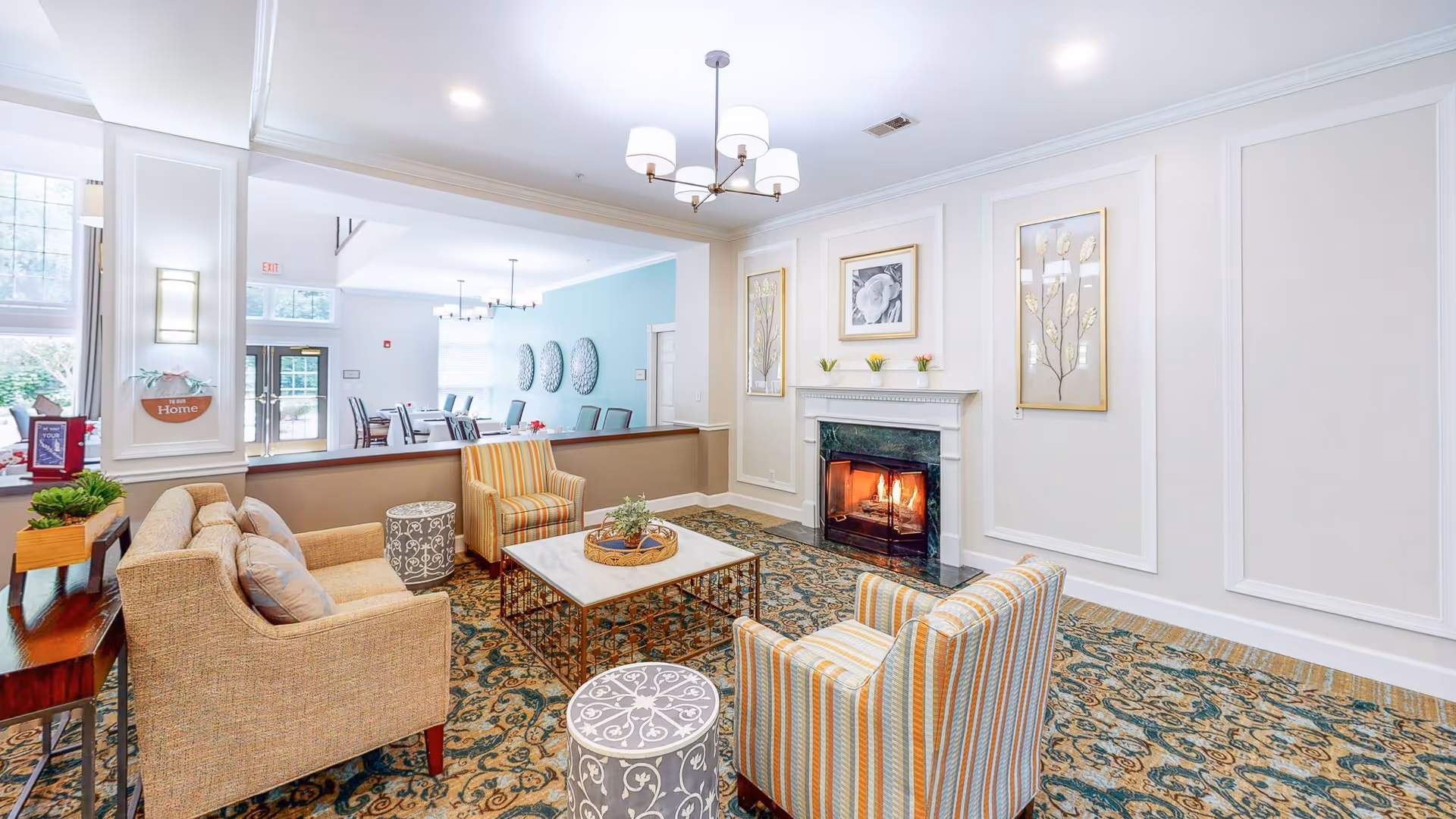 A cozy living room area in a senior living facility with a lit fireplace, a beige sofa, two striped armchairs, a marble coffee table with a decorative tray, and two patterned round stools. The walls are decorated with framed artwork and the room opens into a dining area with tables and chairs. The space is well-lit with ceiling lights and natural light from large windows.