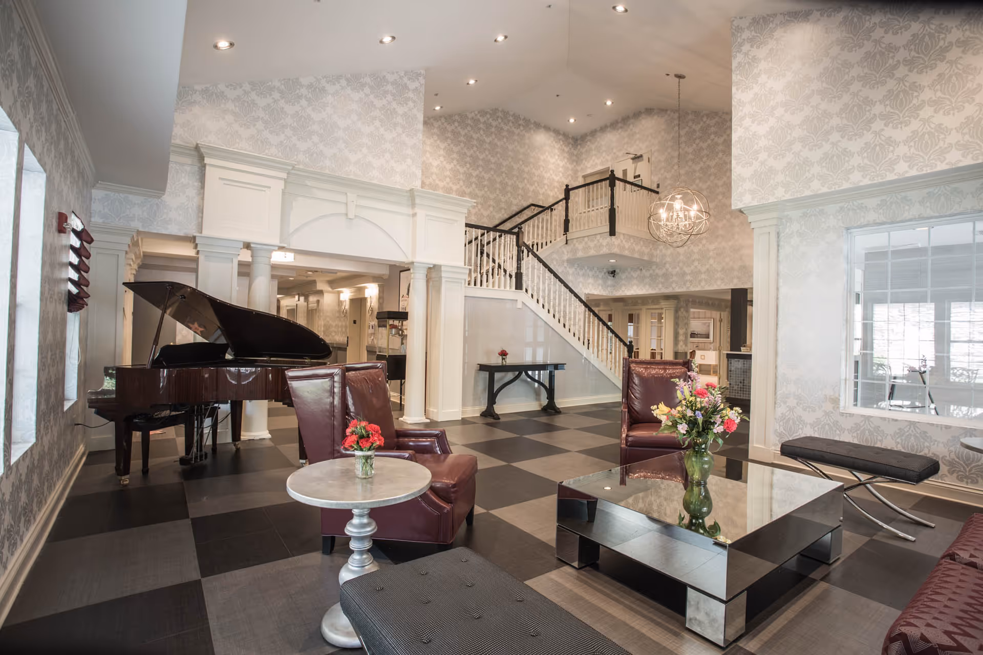 A spacious and elegant assisted living facility common area with a grand piano on the left, two maroon leather armchairs, a mirrored coffee table with a vase of flowers, a small round side table with a flower vase, and a cushioned bench. The room features patterned wallpaper, a staircase with white railings and dark handrails, and a modern chandelier hanging from the ceiling.