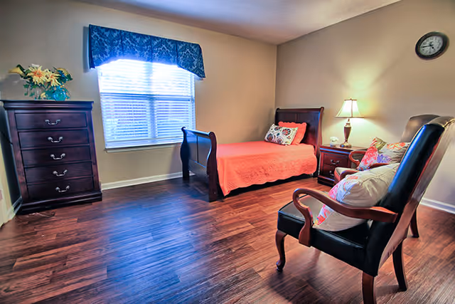 Bright furnished bedroom with a single sleigh bed, wooden dresser, armchair and nightstand on hardwood floors.