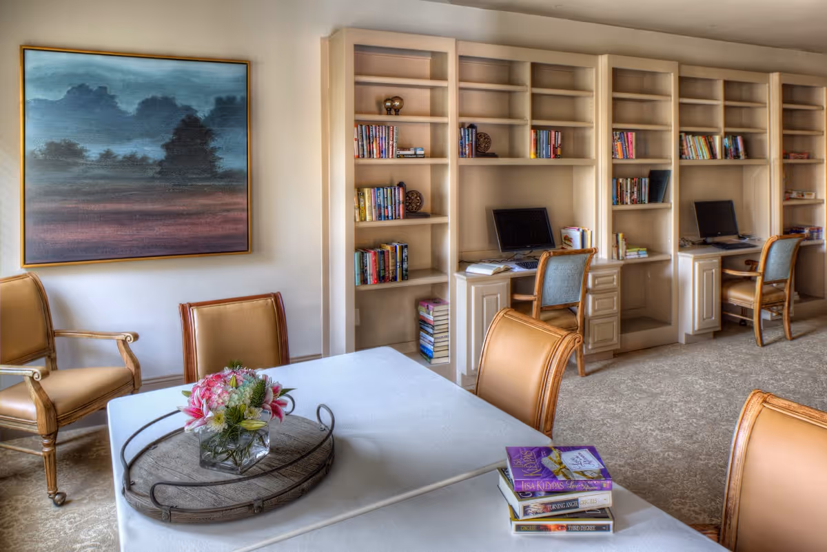 A cozy senior living common area with a white table covered by a cloth, a small flower arrangement in a glass vase on a decorative tray, and a stack of books. Surrounding the table are cushioned wooden chairs. In the background, there are built-in bookshelves filled with books and two computer workstations with chairs. A large framed landscape painting hangs on the wall.