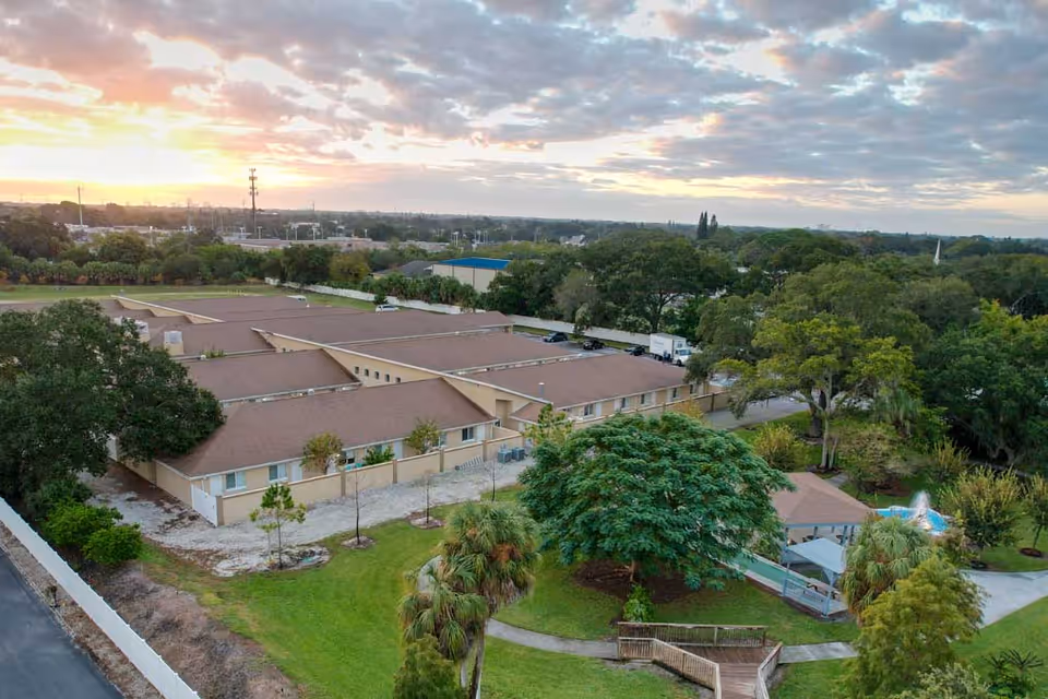 Aerial view of The Goldton at St. Petersburg facility showing a single-story building with a brown roof surrounded by green trees and landscaped grounds. There is a small gazebo and a fountain visible in the garden area, with a partly cloudy sky at sunset in the background.
