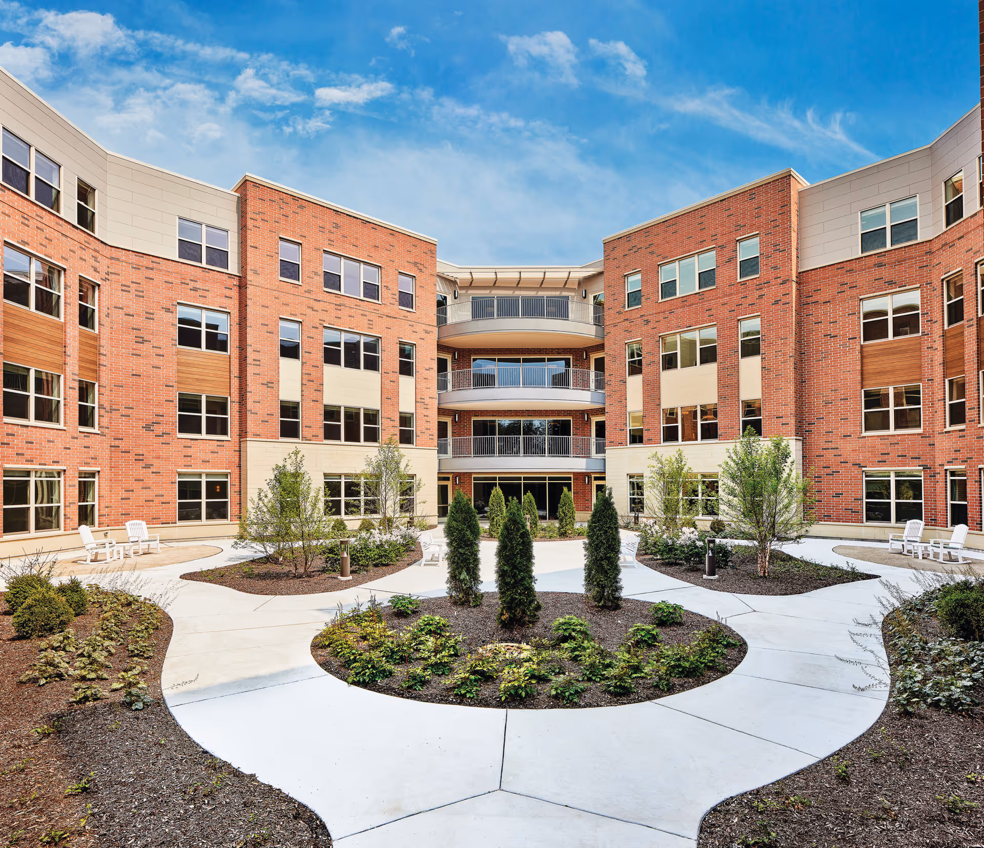 Brick multi-story senior living building surrounding a landscaped central courtyard with curved walkways, small trees, and outdoor seating under a blue sky.