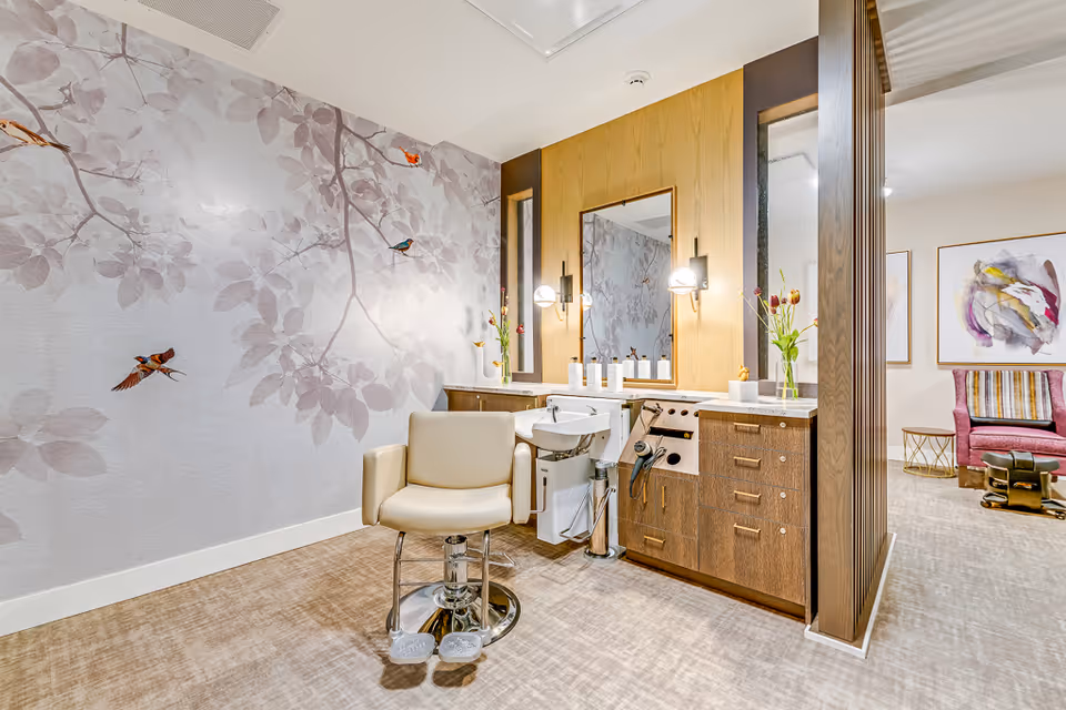 A bright and modern salon area in a senior living facility featuring a beige salon chair in front of a wooden vanity with a large mirror, two wall-mounted lights, and various hair care products. The wall behind the chair has a decorative mural with branches and colorful birds. To the right, there is a glimpse of a cozy seating area with a pink armchair, a striped pillow, and abstract artwork on the wall.