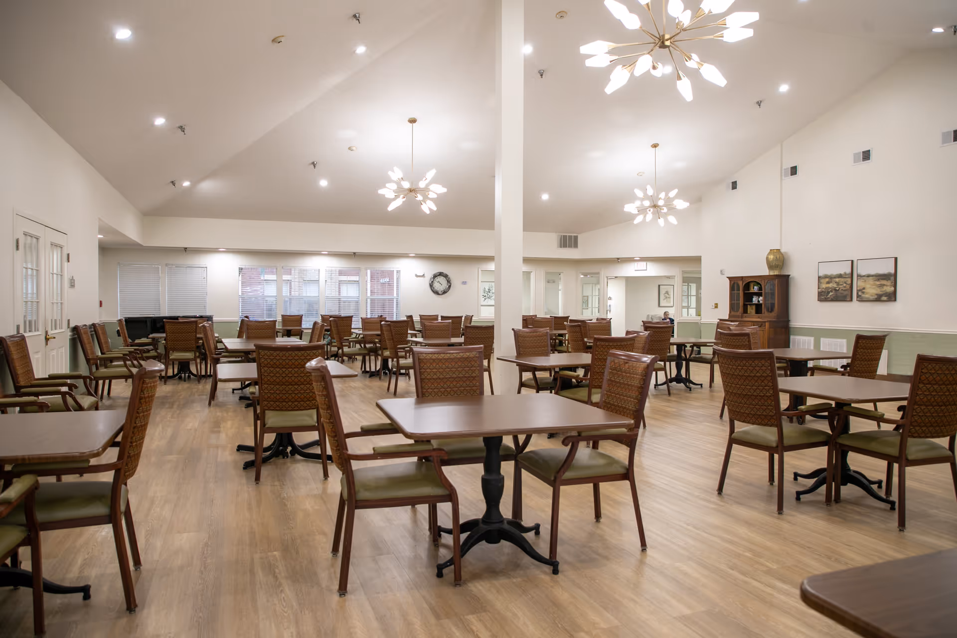 A spacious dining room with multiple wooden tables and chairs arranged neatly on a wooden floor. The room has high ceilings with modern light fixtures, large windows with blinds, and light-colored walls decorated with a clock and framed artwork. There is a wooden cabinet in the corner and a piano near the windows.