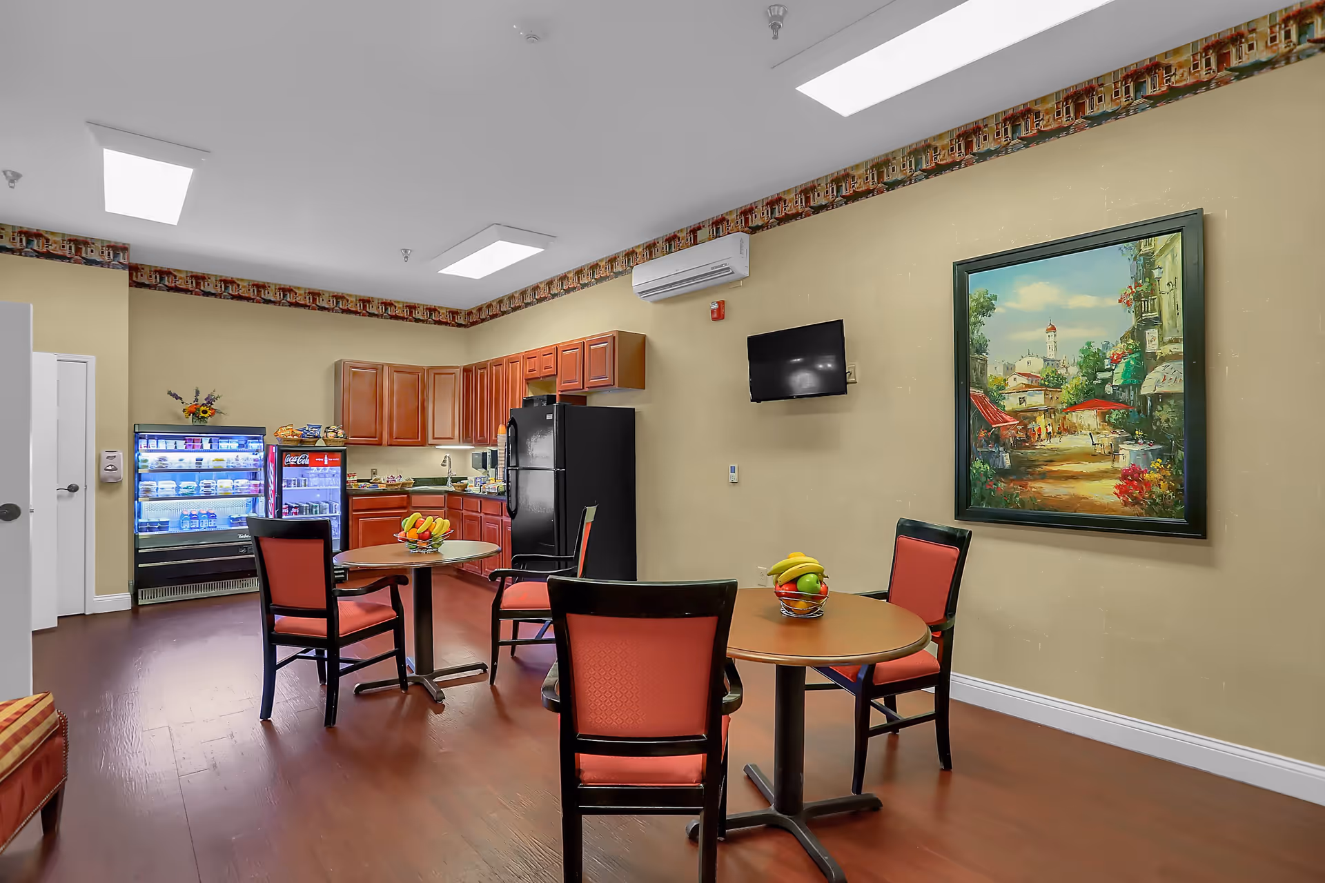 A small kitchen and dining area with wooden cabinets, a black refrigerator, a glass-front beverage cooler, and two round tables each with three chairs. Bowls of fruit are placed on the tables. A colorful painting hangs on the beige wall, and a small flat-screen TV is mounted nearby. The room has hardwood floors and decorative wallpaper border near the ceiling.